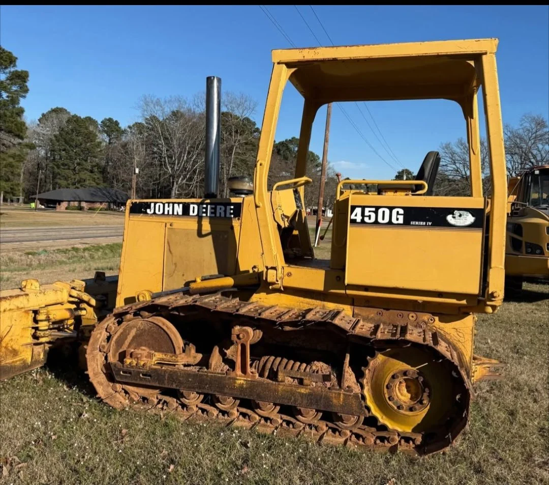 A yellow John Deere 450G Series IV bulldozer with rust on the tracks, parked on grass near a road with trees and a small building in the background.