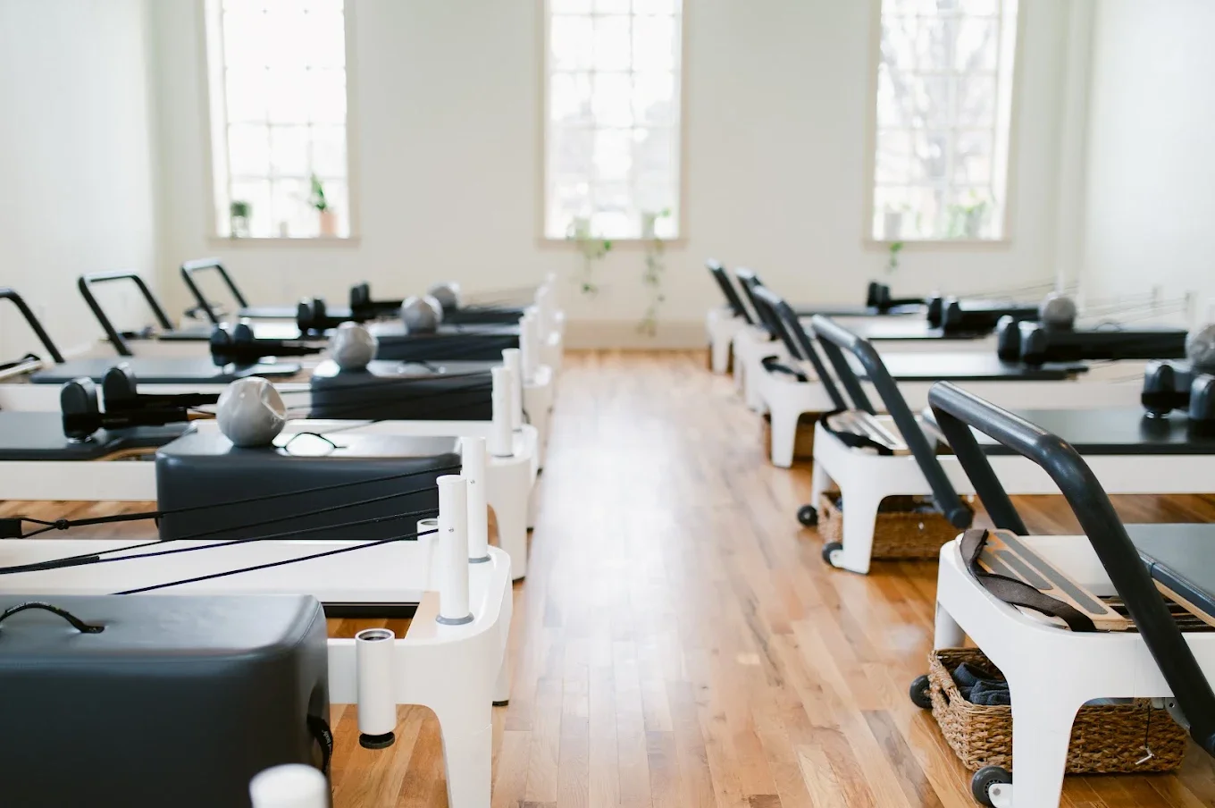 Row of Pilates reformer machines in a bright, spacious fitness studio with wooden floors and large windows.