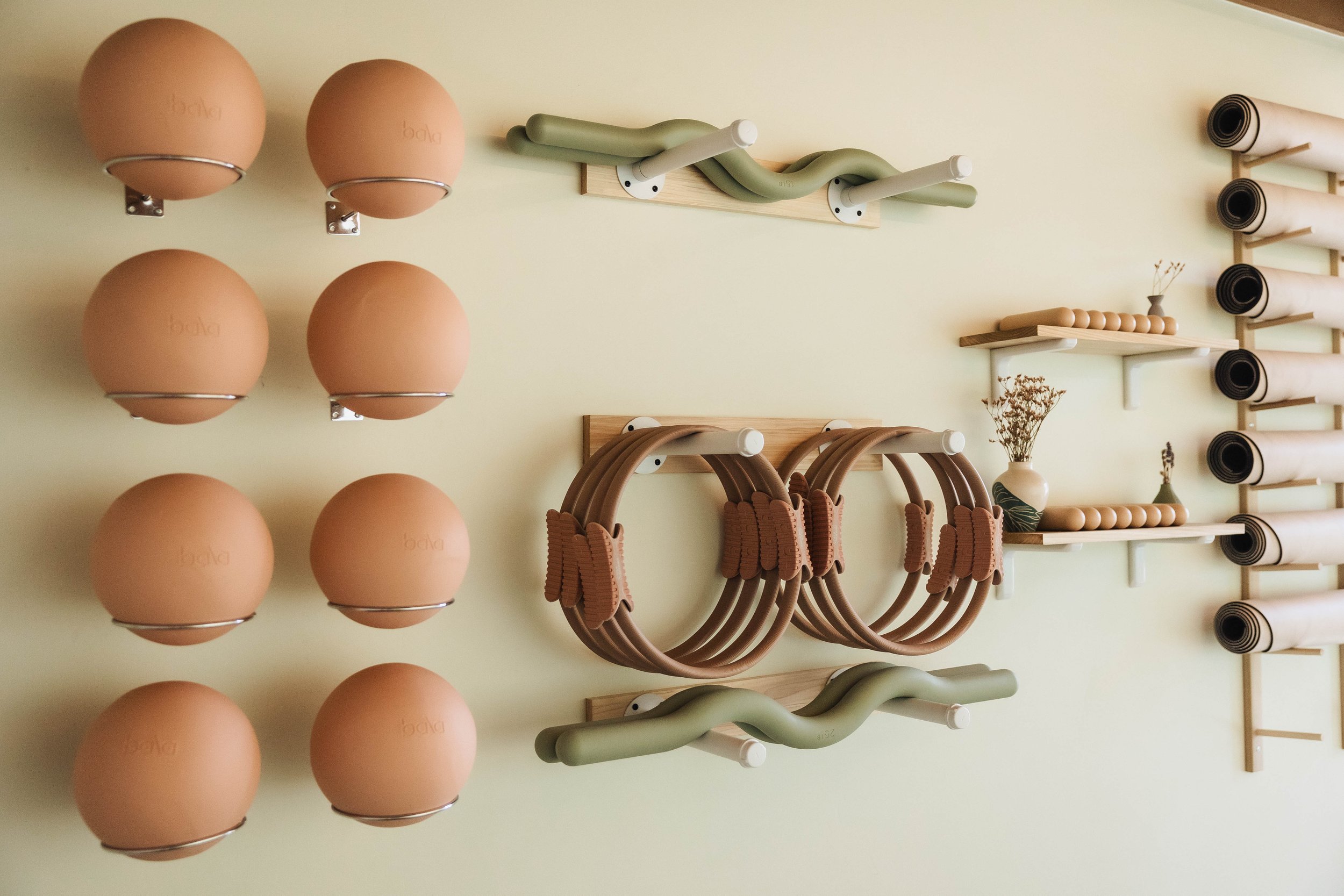 Display of various fitness equipment including peach-colored exercise balls, green and beige resistance bands, and wooden shelves with rolled mats and bean bags against a light-colored wall.