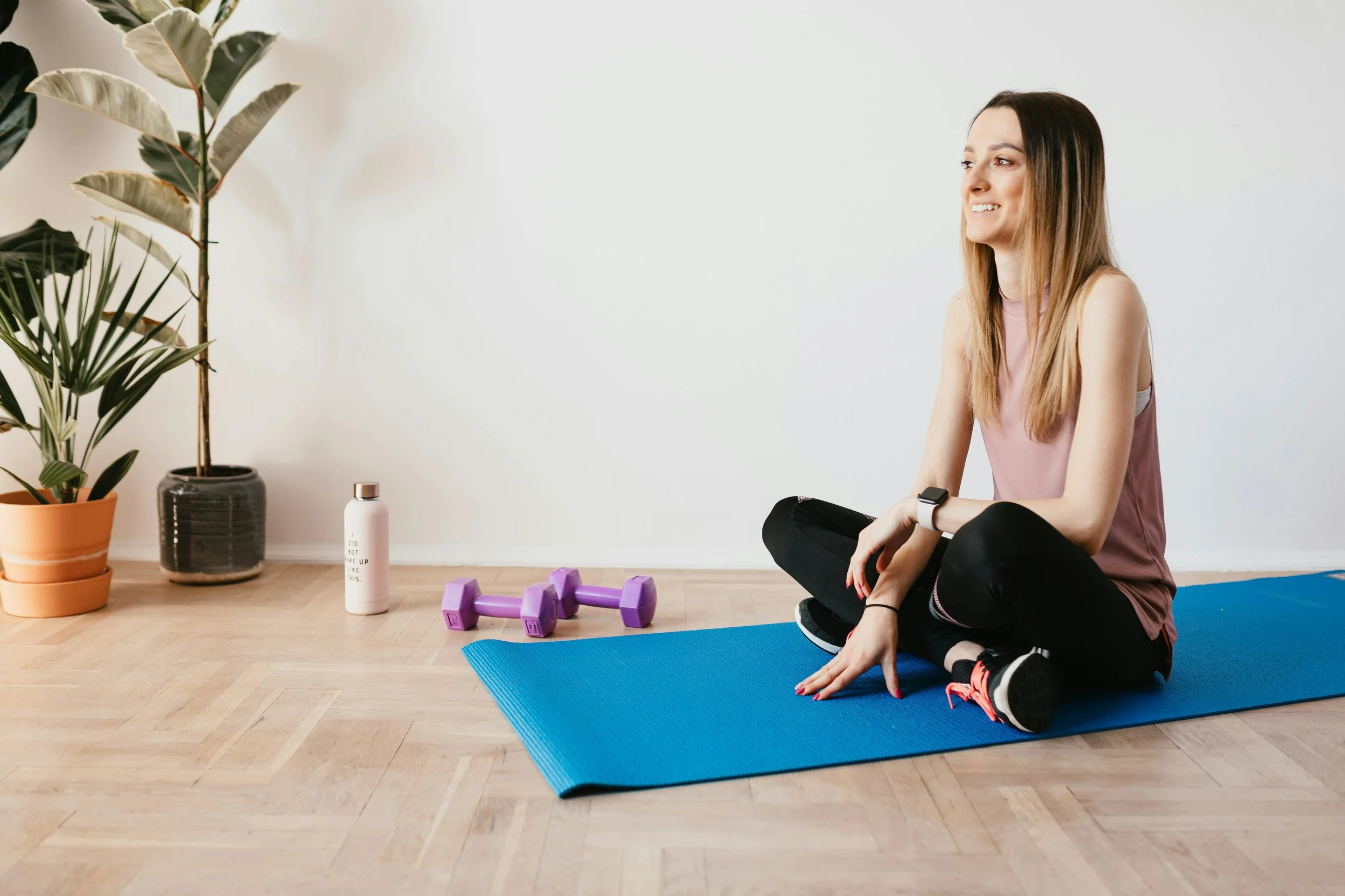A woman sitting cross-legged on a blue exercise mat, smiling while resting her right arm on her knee, in a room with wooden floors and white walls. Next to her are light purple dumbbells. Behind her are potted plants and a white bottle.