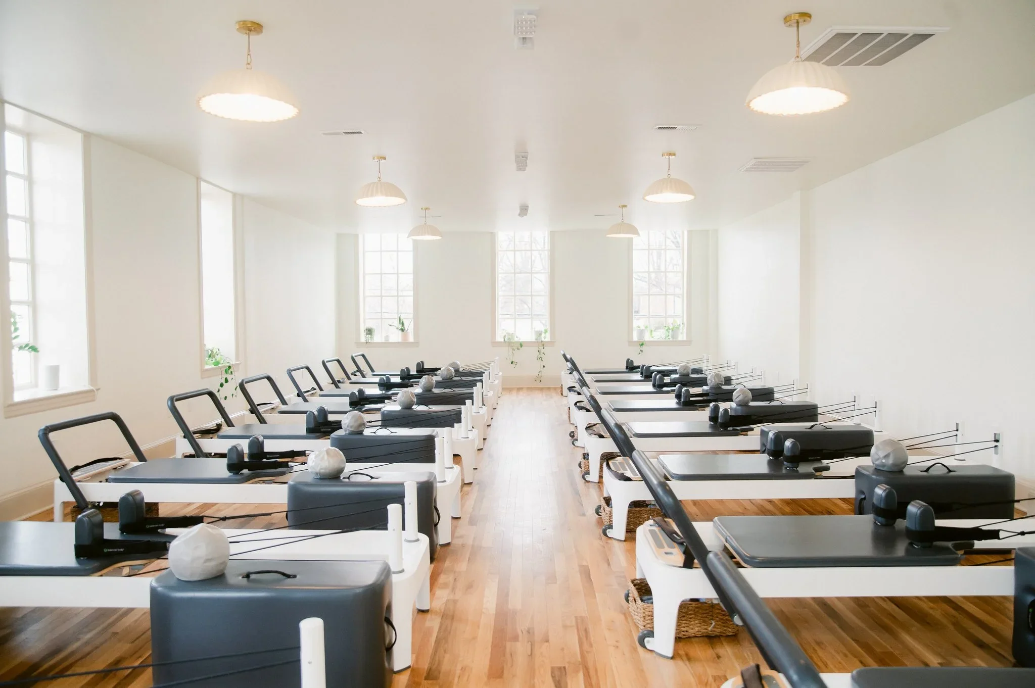 A room with multiple Pilates reformer machines arranged in rows, each with white pillows, in a well-lit room with large windows and wooden floors.