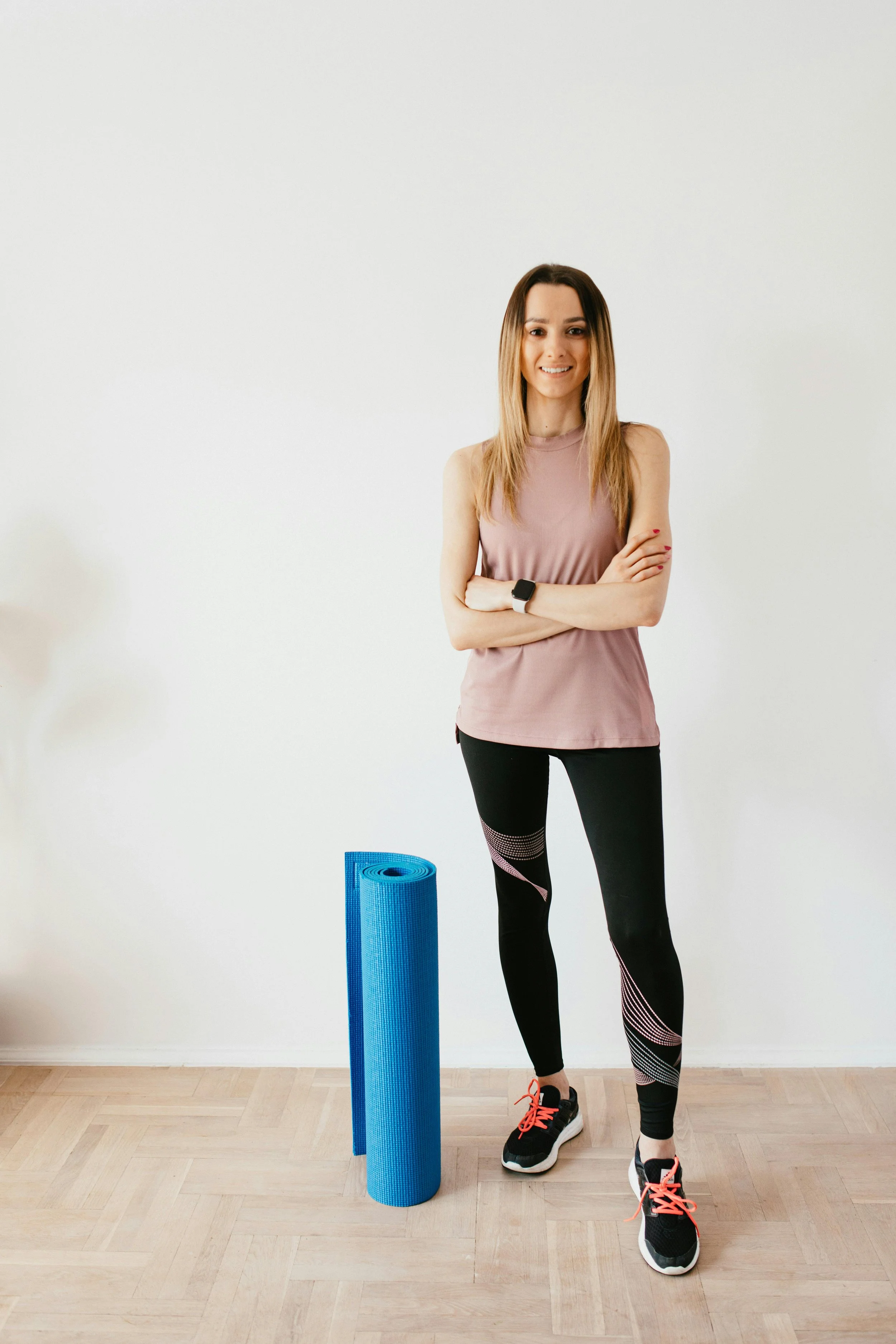 A woman standing indoors on a wooden floor, dressed in athletic wear, smiling, with arms crossed. A rolled-up blue yoga mat is on the floor beside her.