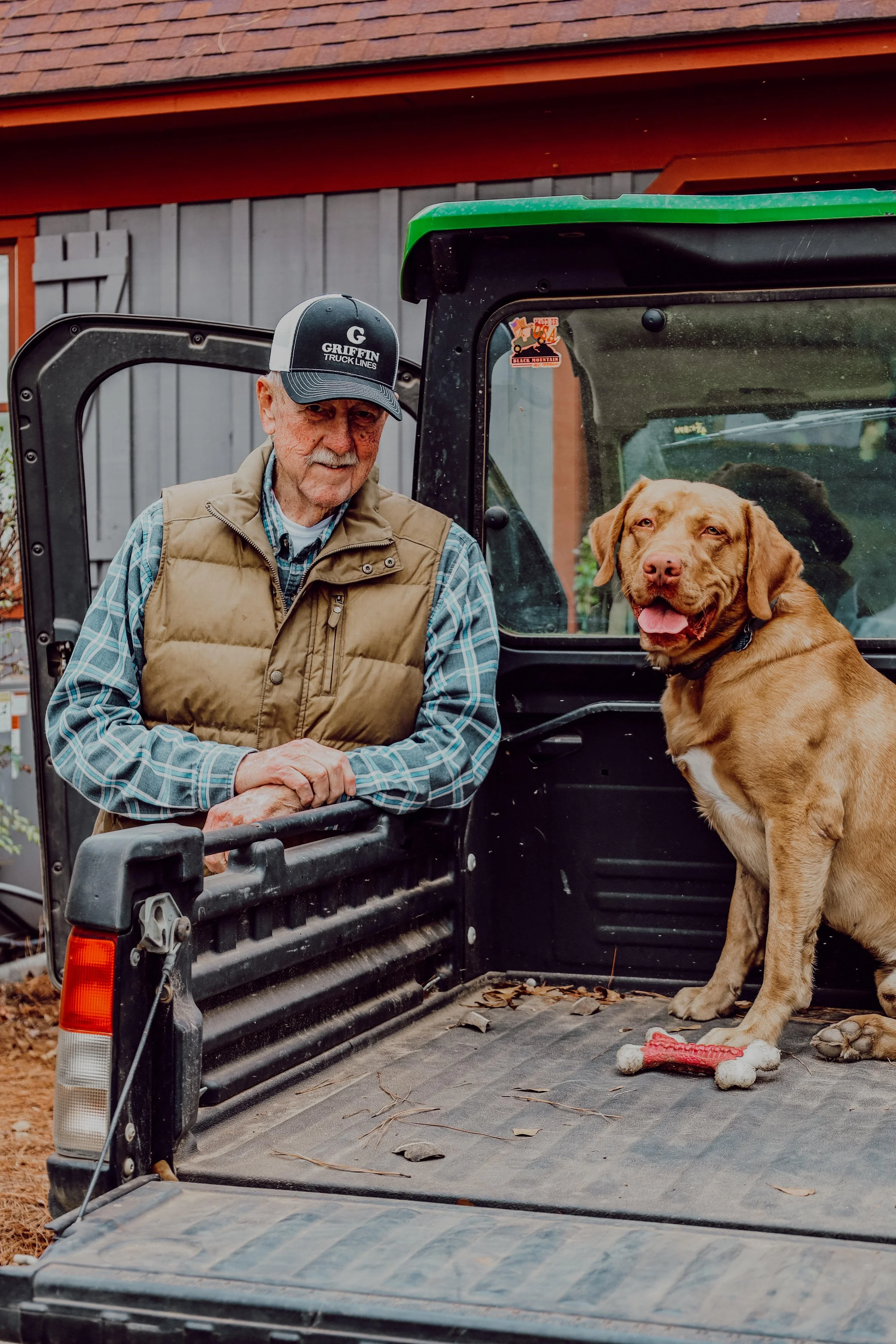 An older man in a plaid shirt and beige vest leaning on the side of a black utility vehicle, with a yellow Labrador Retriever sitting in the truck bed next to a dog toy, outside a building with wooden siding.