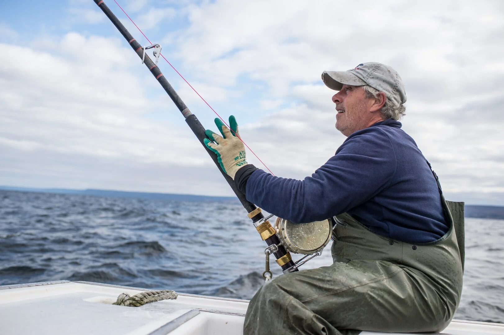 Man fishing on a boat in the ocean, wearing gloves, a blue sweatshirt, green waterproof overalls, and a gray cap.