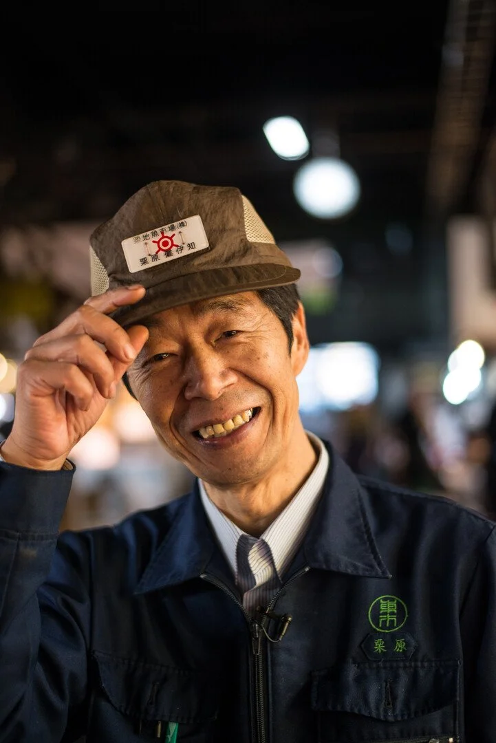 Smiling man in dark jacket adjusting cap in indoor market or restaurant setting.