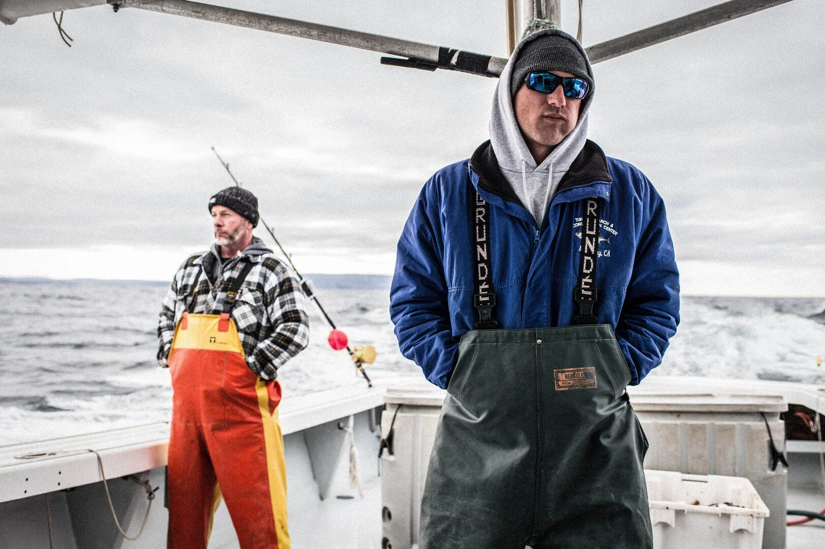 Two fishermen on a boat, one in a blue jacket and sunglasses standing in the foreground, the other in a plaid shirt and orange and yellow waterproofs fishing in the background, on a cloudy day at sea.