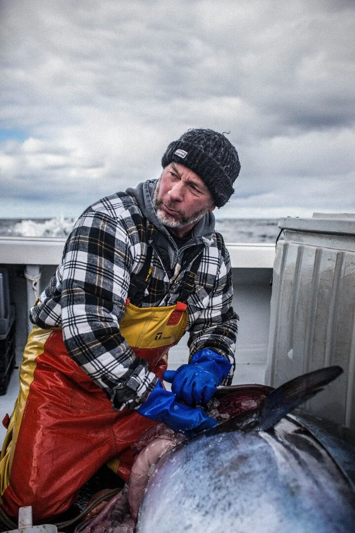An man in a plaid shirt, black beanie, and yellow waterproof apron is on a boat, handling a large fish against a cloudy sky.