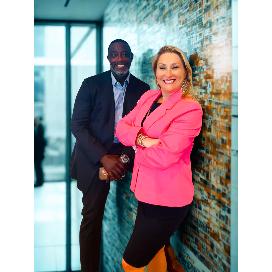 A woman in a pink blazer and black skirt, smiling with arms crossed, standing next to a man in a dark suit, both posing in front of a textured wall.
