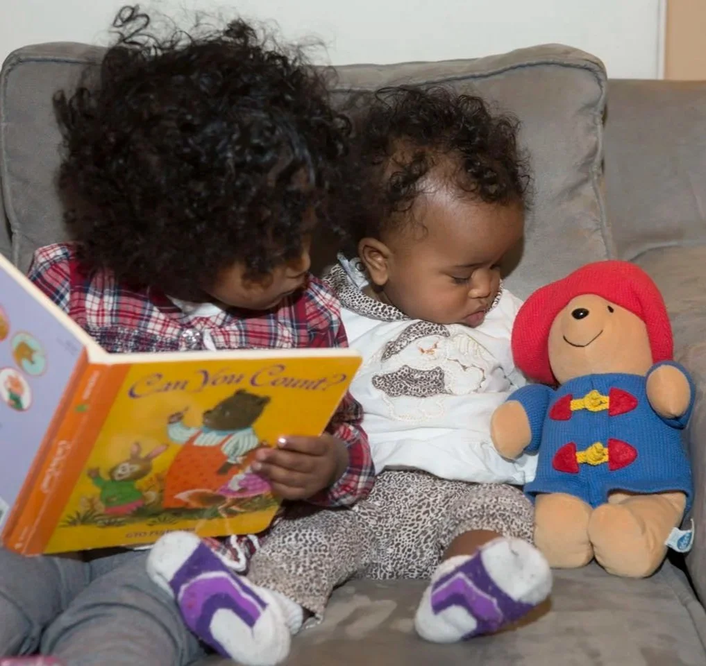 Two young children sitting together on a couch, one reading a children's book titled 'Can You Count?', the other looking at a plush doll of a character with a red hat and blue outfit.