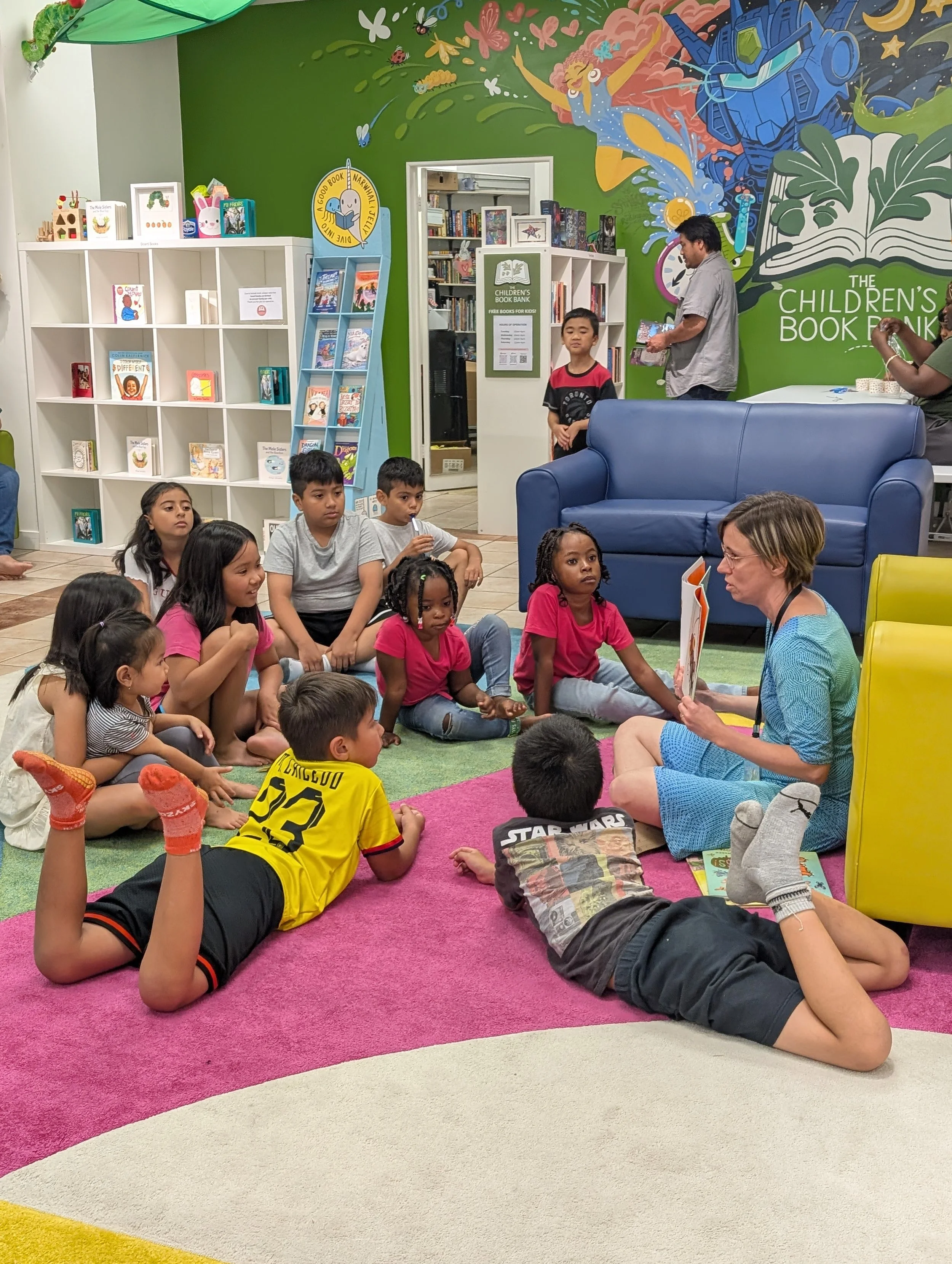 A woman reads a children's book to a group of children sitting on a pink and white carpet in a library or bookstore. The background features bookshelves, a colorful mural, and signs indicating a children's book bank.