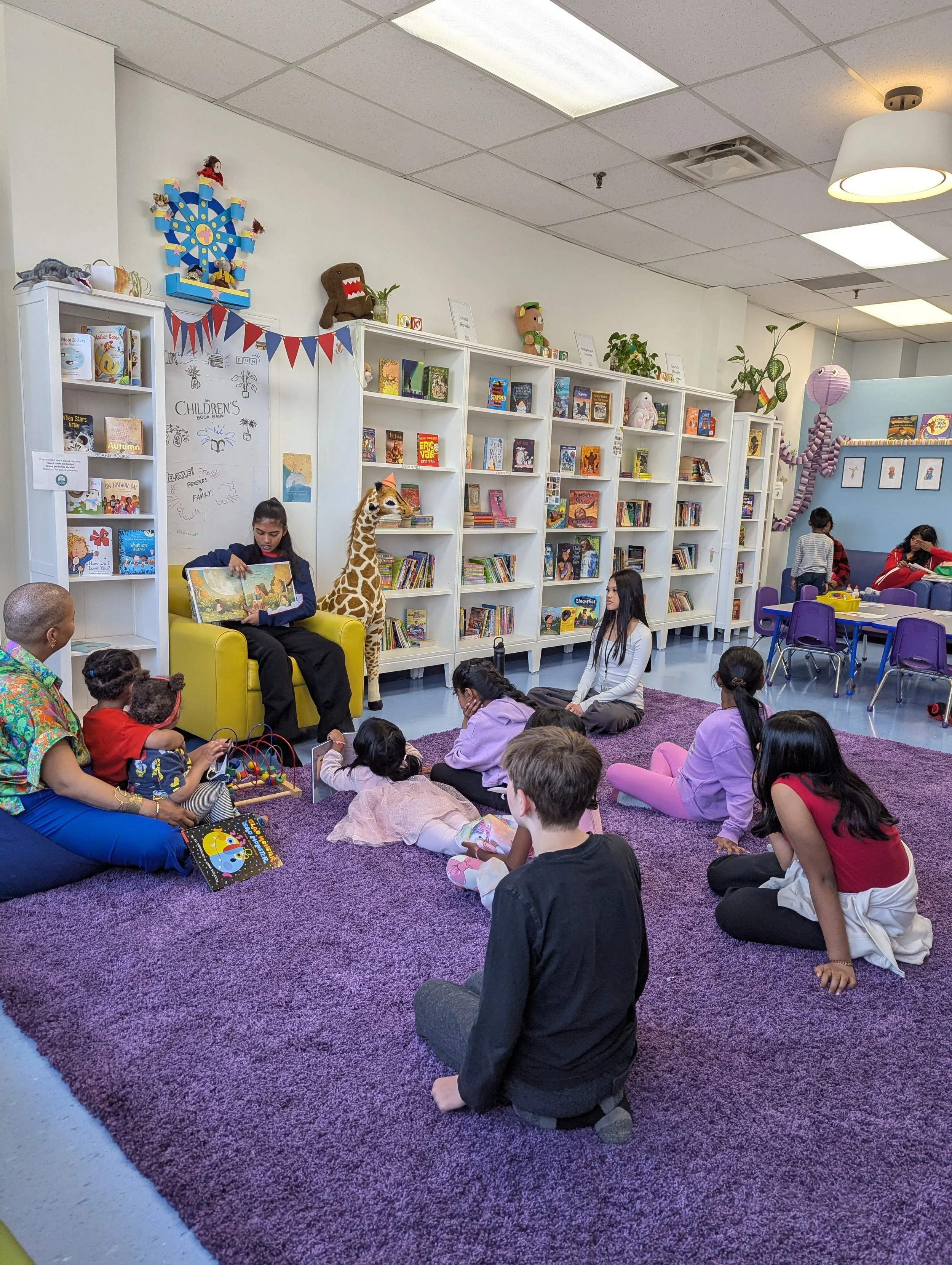 Children sitting on a purple carpet, listening to a story being read by a woman in a library or bookstore. Shelves filled with books and plush toys, with colorful decorations and a large stuffed giraffe.