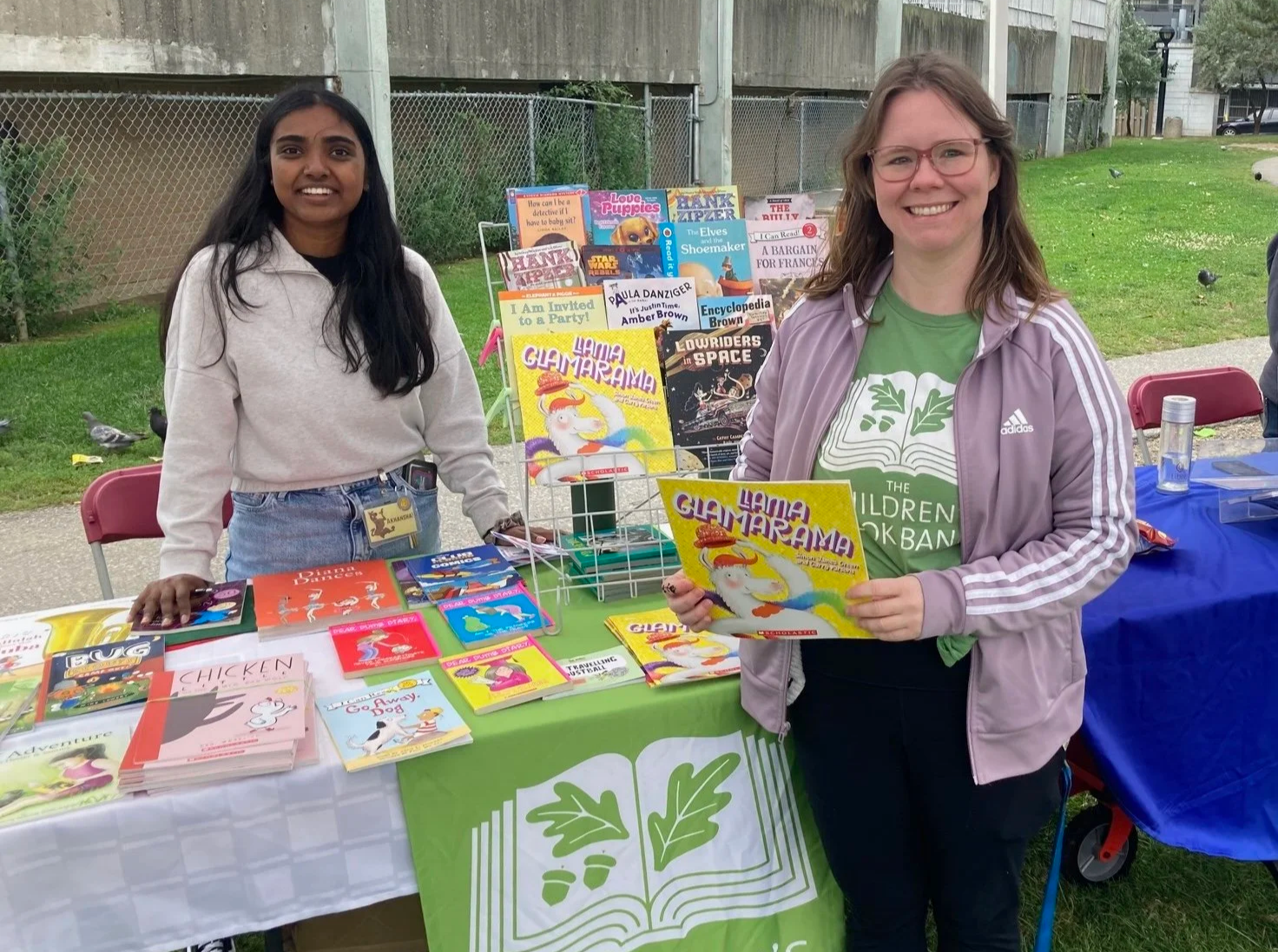 Two Book Bank staff standing at a table outdoors, with one woman holding a colourful children's book. The table is filled with children's books and a book display stand behind them. They are smiling, and the setting appears to be a community event