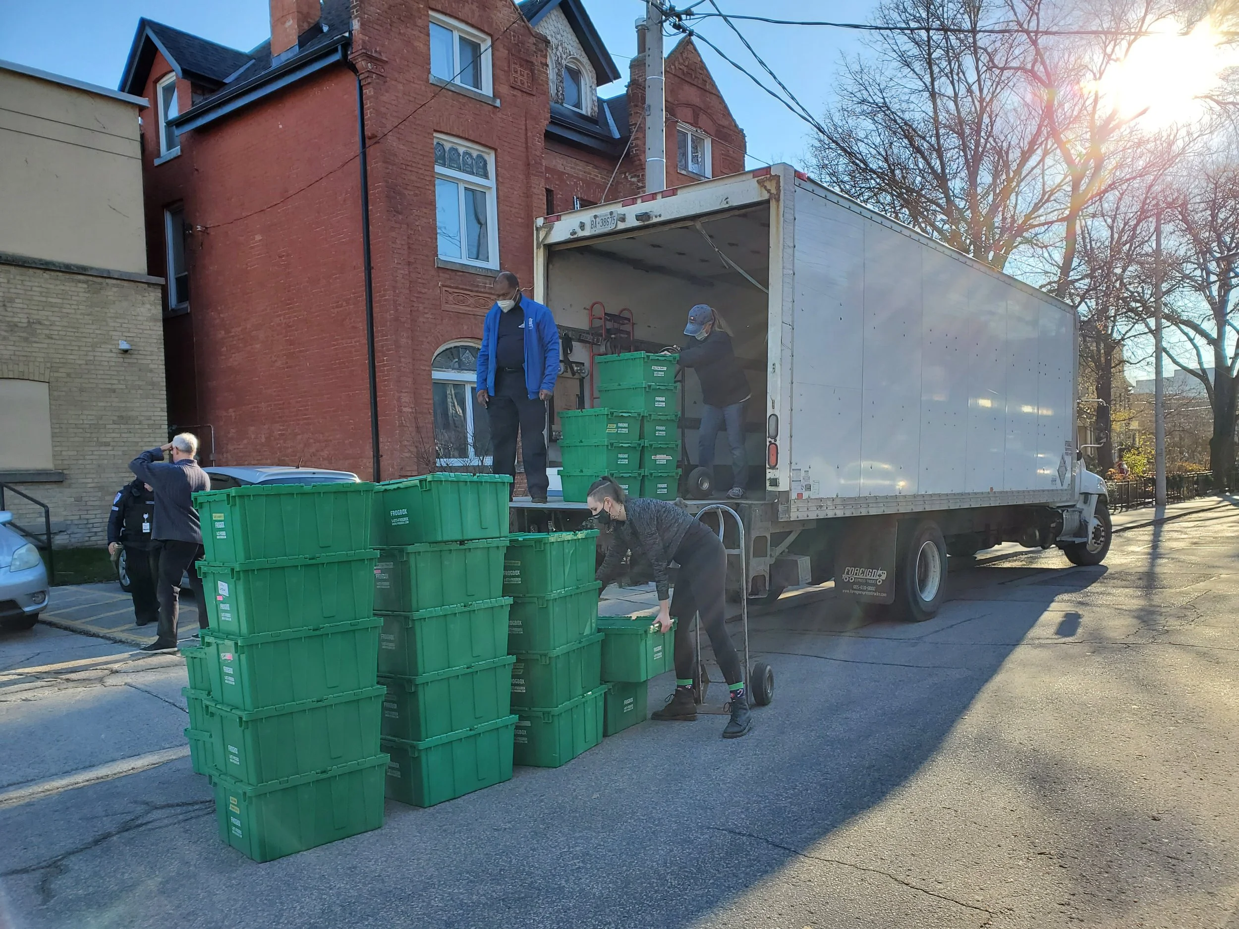 People unloading green bins from a white moving truck on a city street on a sunny day, with a red brick building and leafless trees in the background.