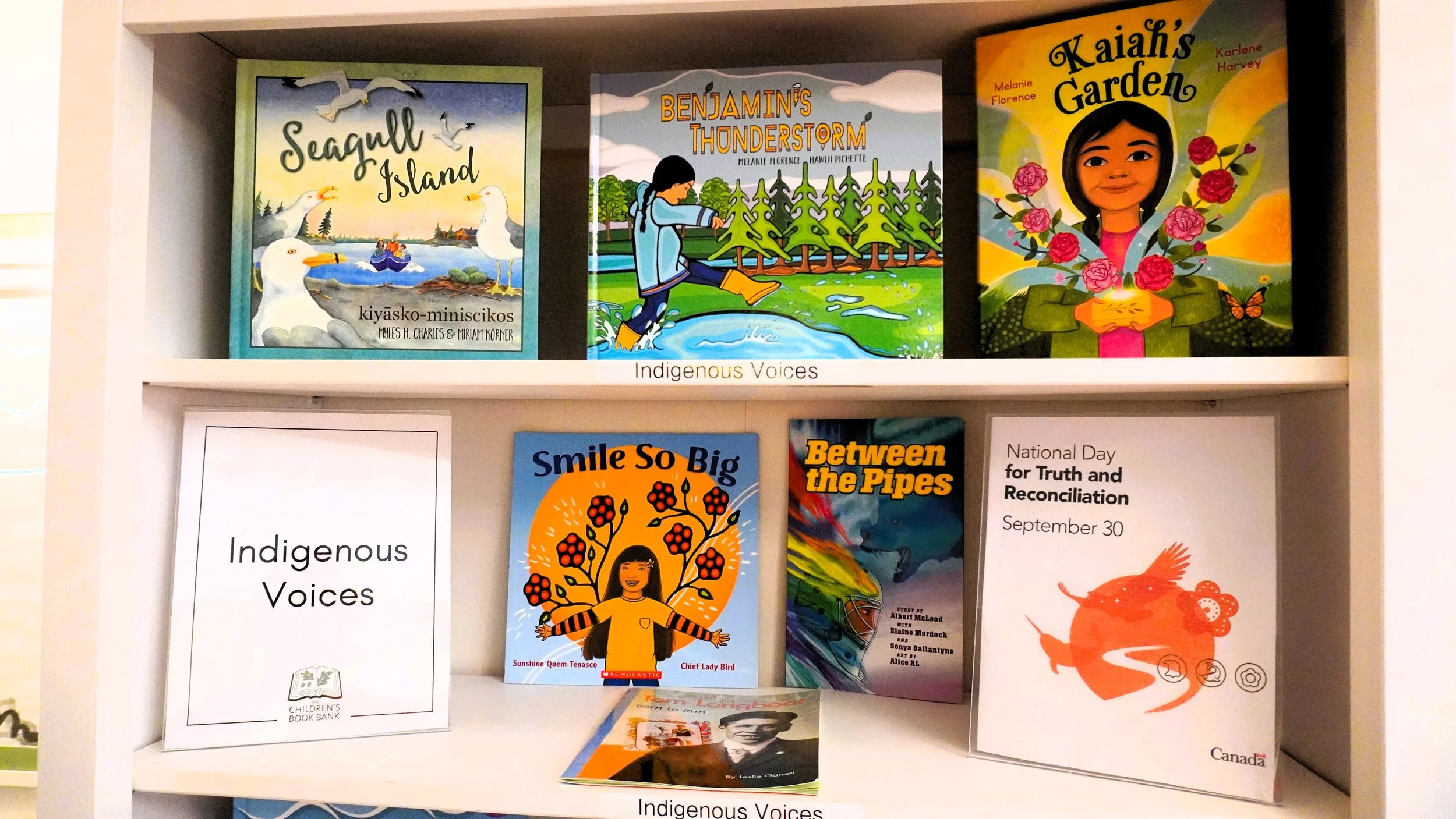 A display shelf with books about Indigenous voices and stories, including titles like 'Seagull Island,' 'Benjamin's Thundersorm,' 'Kaia's Garden,' 'Smile So Big,' 'Between the Pipes,' and posters for National Day for Truth and Reconciliation.