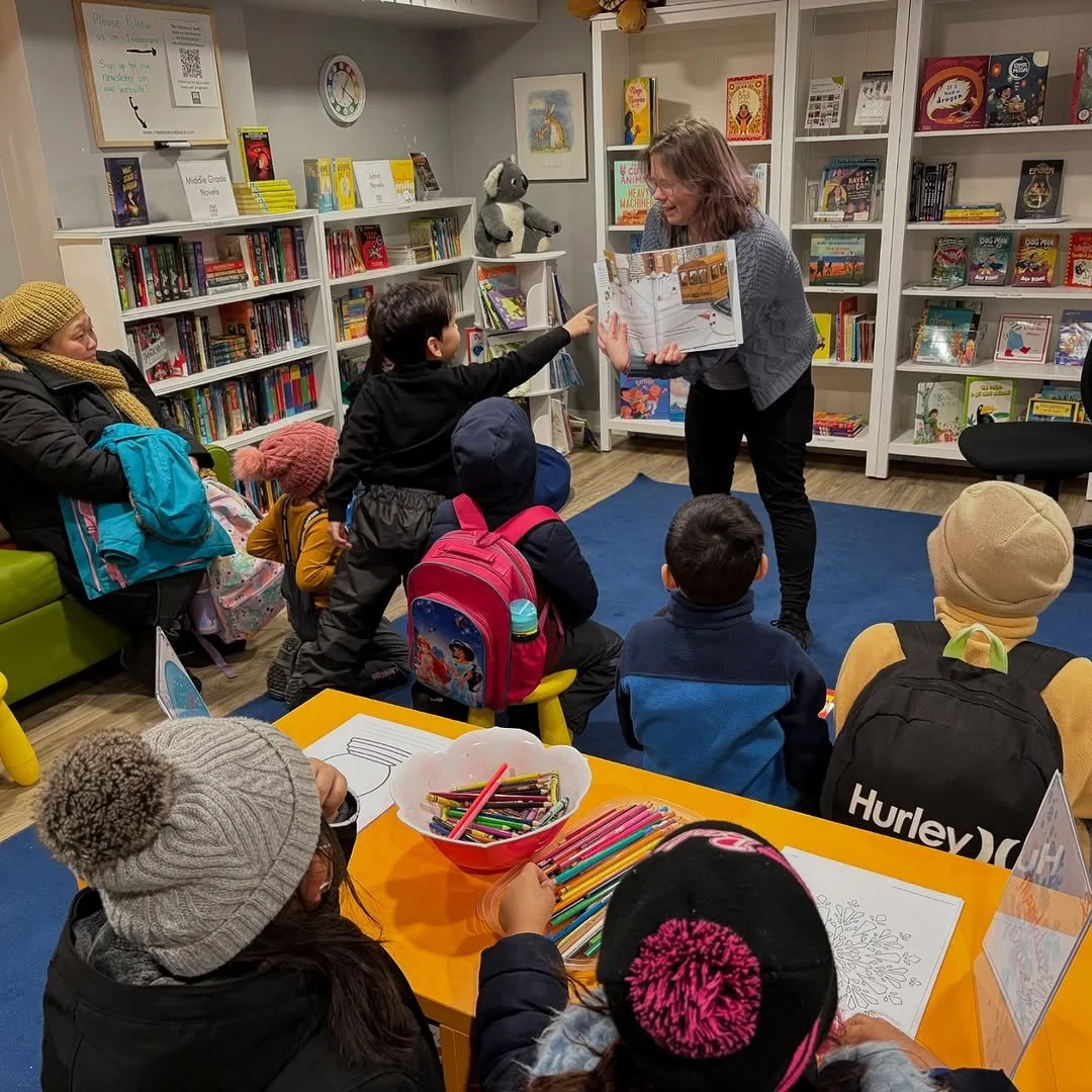 A woman reads a children's book aloud to a group of children in a bookstore or library, with some children sitting on the floor and others at a table with coloring supplies.