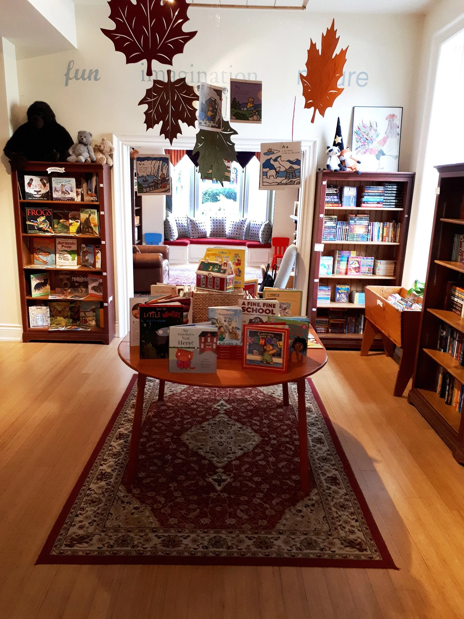 A cozy library room with a wooden oval table in the center displaying children's books. The room has bookshelves filled with books, stuffed animals, and decorative leaves hanging from the ceiling. There is a reading nook with pillows near a window in the background.