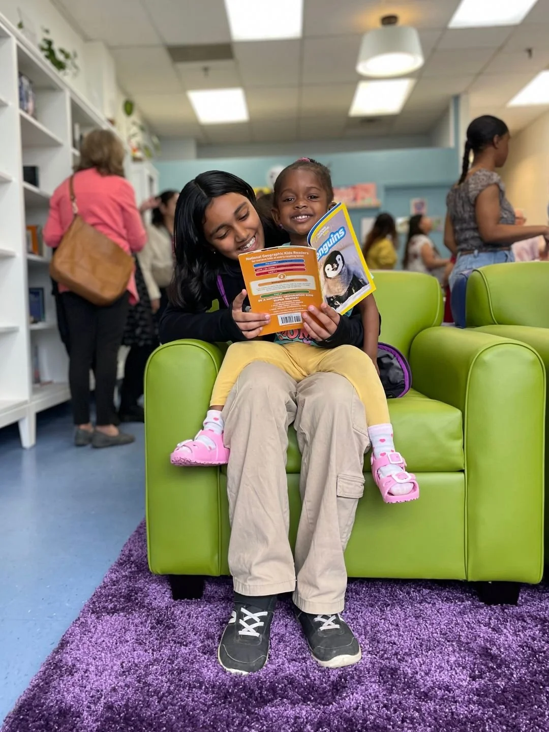 A woman and a young girl sitting on a bright green armchair in a library, smiling and reading children's books together. The girl is wearing pink shoes, yellow pants, and a dark top, while the woman wears beige pants and sneakers. People are visible in the background browsing the shelves.