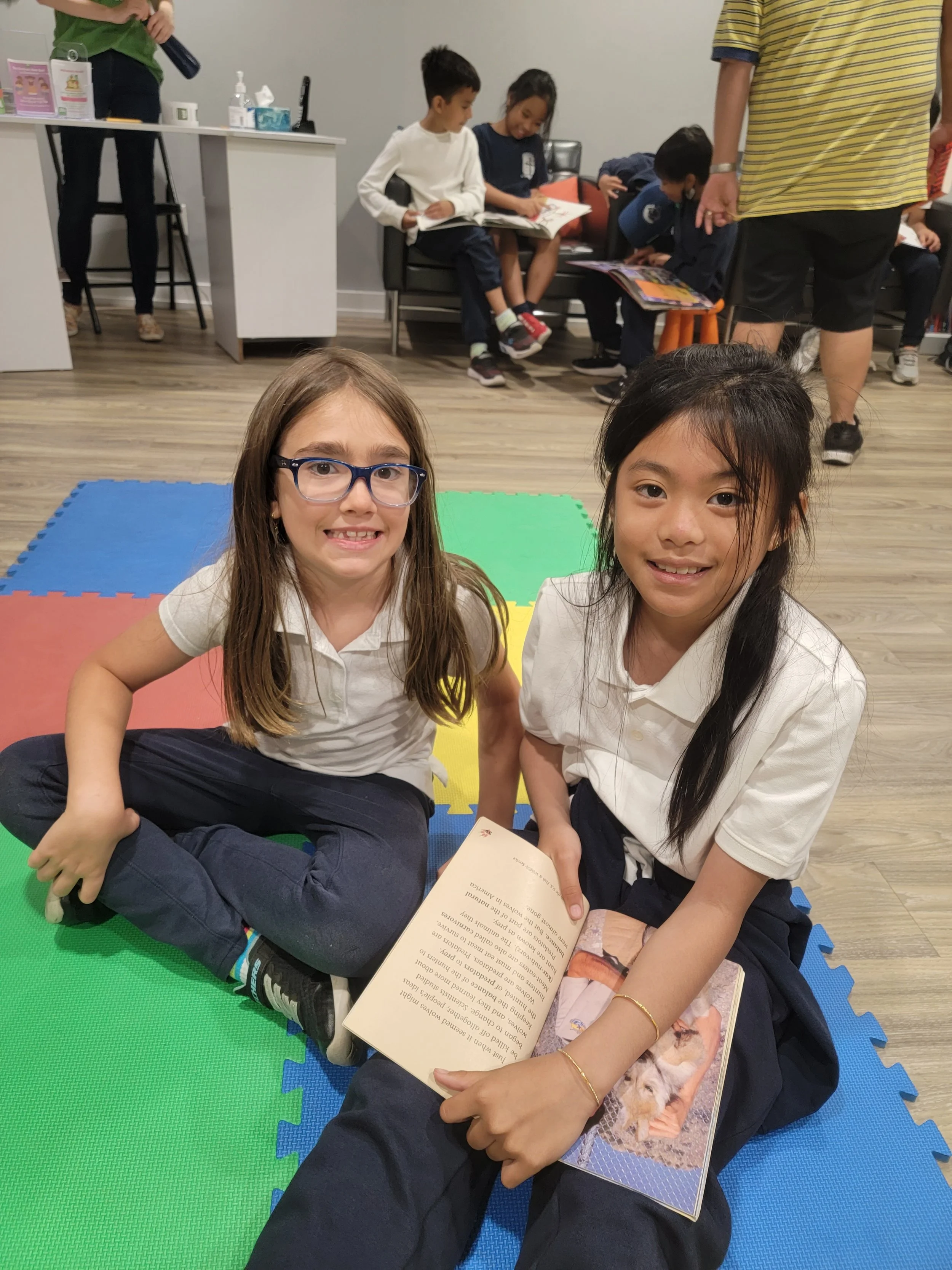 Two young girls in school uniforms sitting on colorful foam mats, reading a book together inside a classroom or library.