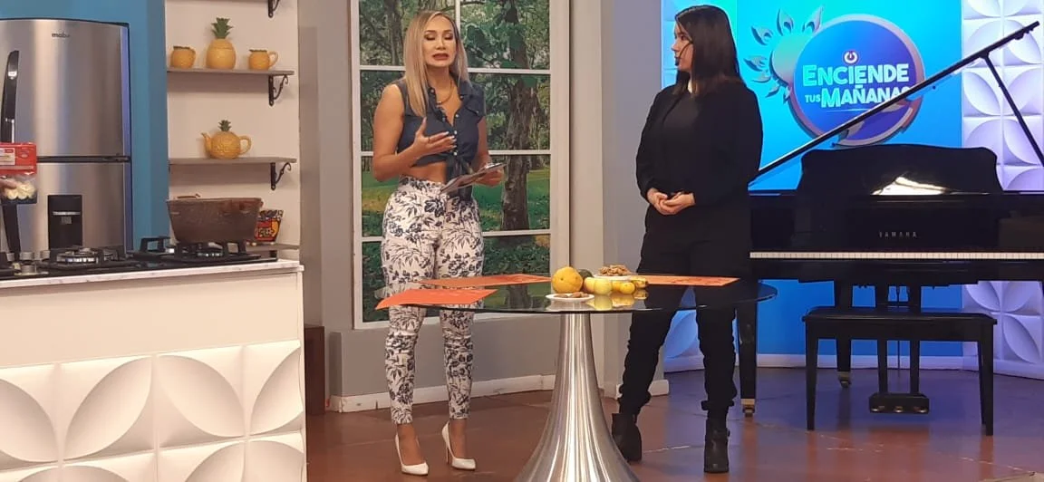 Dos mujeres conversando en un programa de televisión en un set que tiene una mesa redonda, frutas en la mesa y un piano en el fondo.