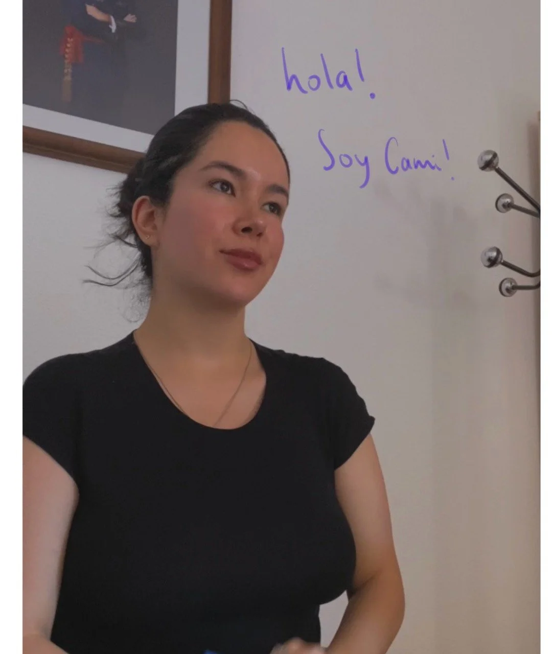 psicologa en quito, oficina al norte, peinado hacia atrás, con camiseta negra, en una sala con paredes blancas. En la pared, hay un cuadro y una pizarra blanca con la inscripción en marcador morado