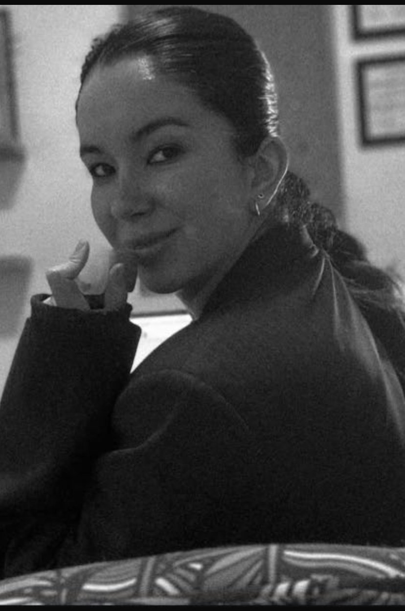 Retrato en blanco y negro de una mujer con cabello recogido, sonriendo y mirando hacia la cámara, con un fondo de cuadros en la pared.