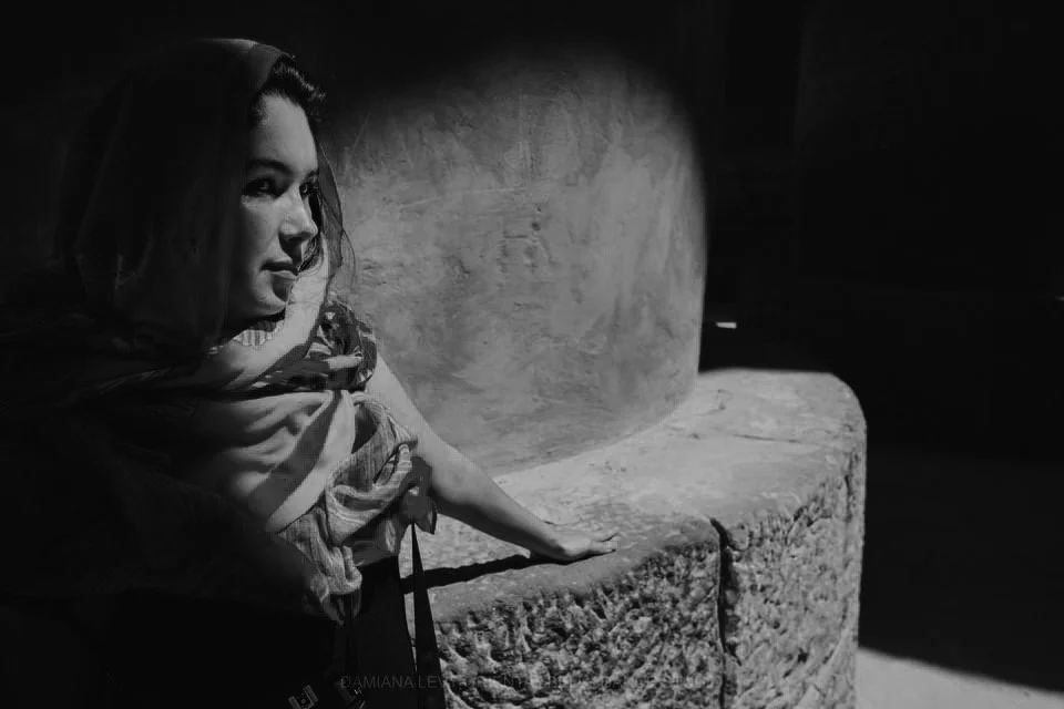 Mujer joven con bufanda en el cuello, recargada sobre una superficie de piedra, en un ambiente en penumbra, en blanco y negro.