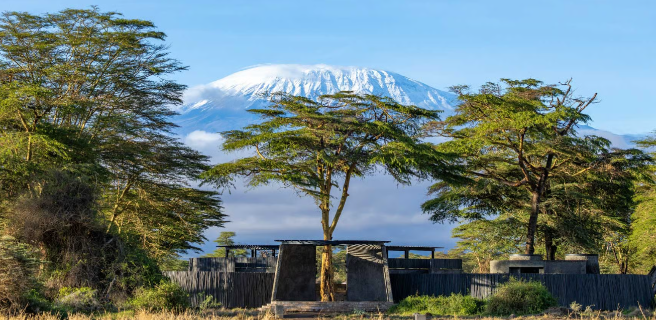 Mount Kilimanjaro snow-capped in the background, with green trees and a black fence and structures in the foreground.