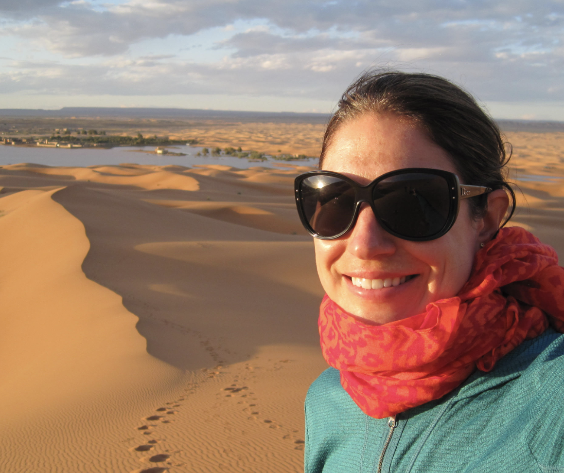 Meredith Fenn wearing sunglasses and a red scarf taking a selfie in a desert with sand dunes of Morocco and a distant body of water in the background.