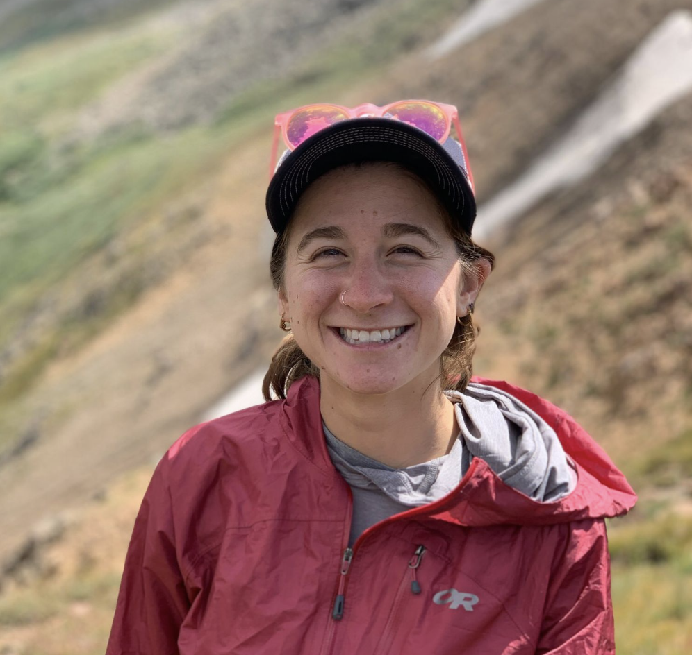 Dru Falco outdoors on a mountain trail, smiling, wearing a red jacket, gray shirt, black baseball cap, and pink sunglasses on her head.