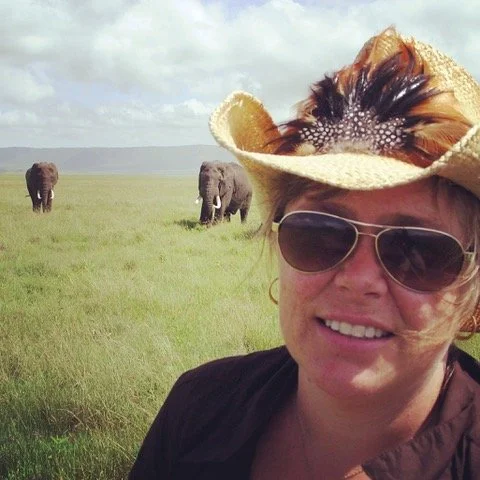 A woman wearing sunglasses and a straw hat with feathers smiling in a grassy savannah, with elephants in the background.