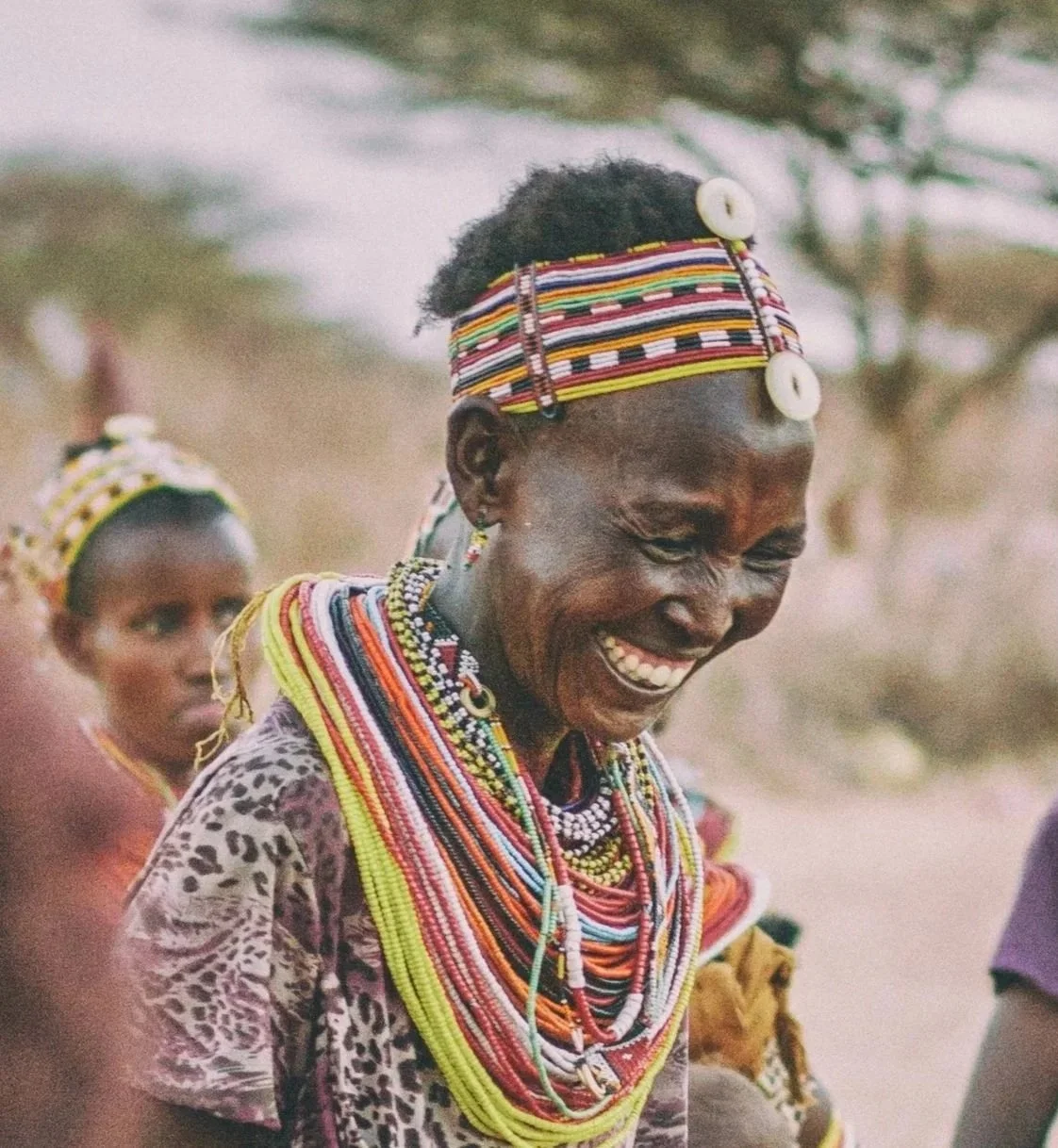Smiling woman from an African tribe wearing colorful beaded jewelry and a patterned headband, with another woman in the background also wearing traditional attire.