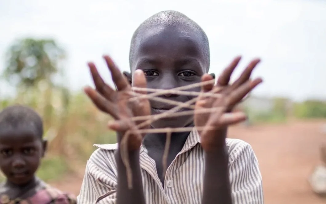 A young boy with a shaved head holding a handmade slingshot made of elastic and sticks, standing outdoors on a dirt path with another boy in the background.