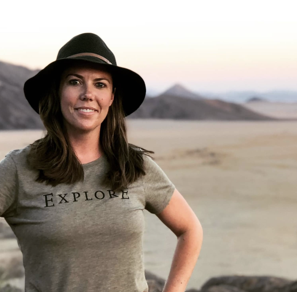 Katie McDonough with long brown hair wearing a gray t-shirt that says 'EXPLORE' and a large black sun hat, standing outdoors in a desert landscape with mountains in the background at sunset.