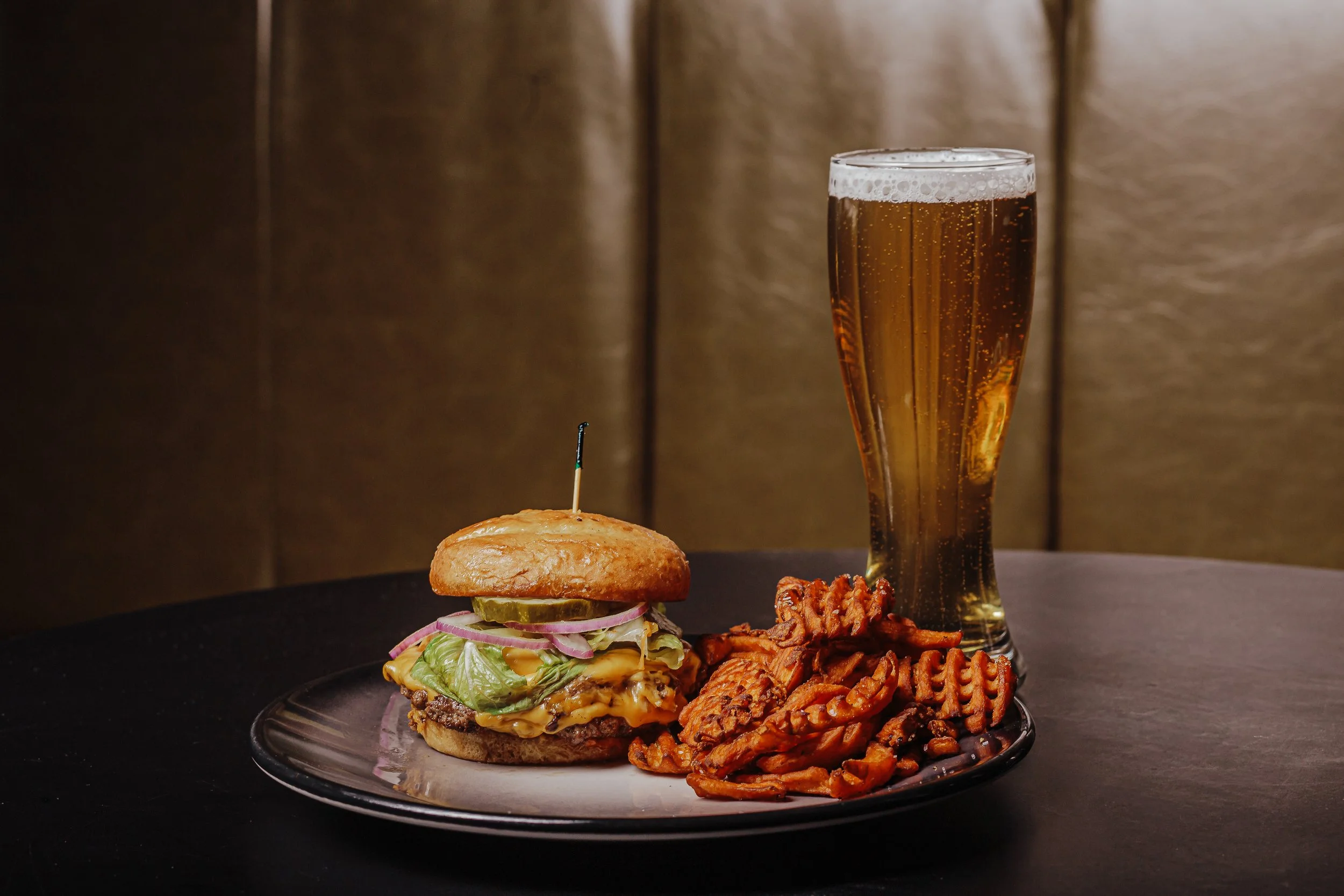 Photo of a smash burger with three patties, onion, lettuce, pickle on a toasty bun with waffle cut sweet potato fries and a light bubbly beer
