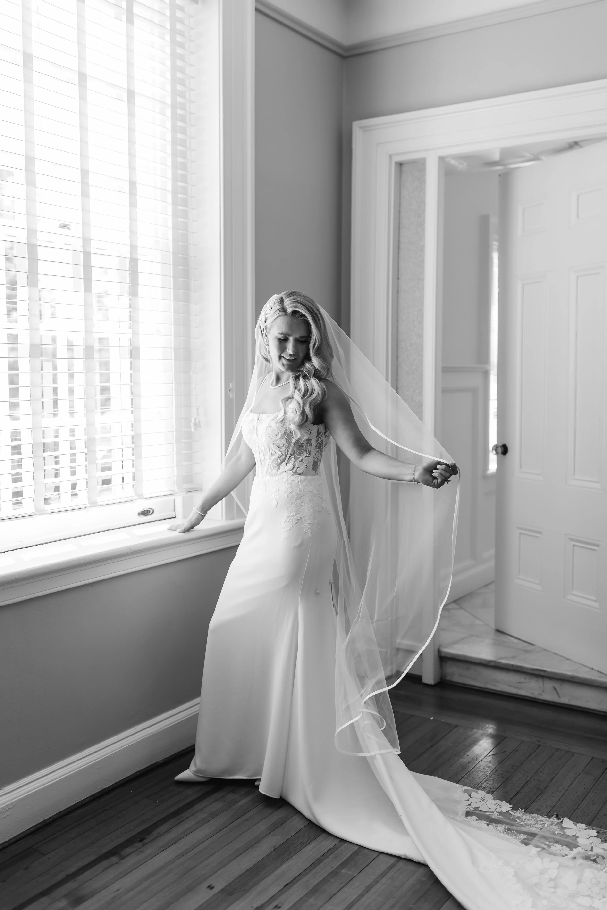 Black and white photo of a bride in a wedding dress with a veil, standing indoors next to a window with blinds, looking down and holding the veil.