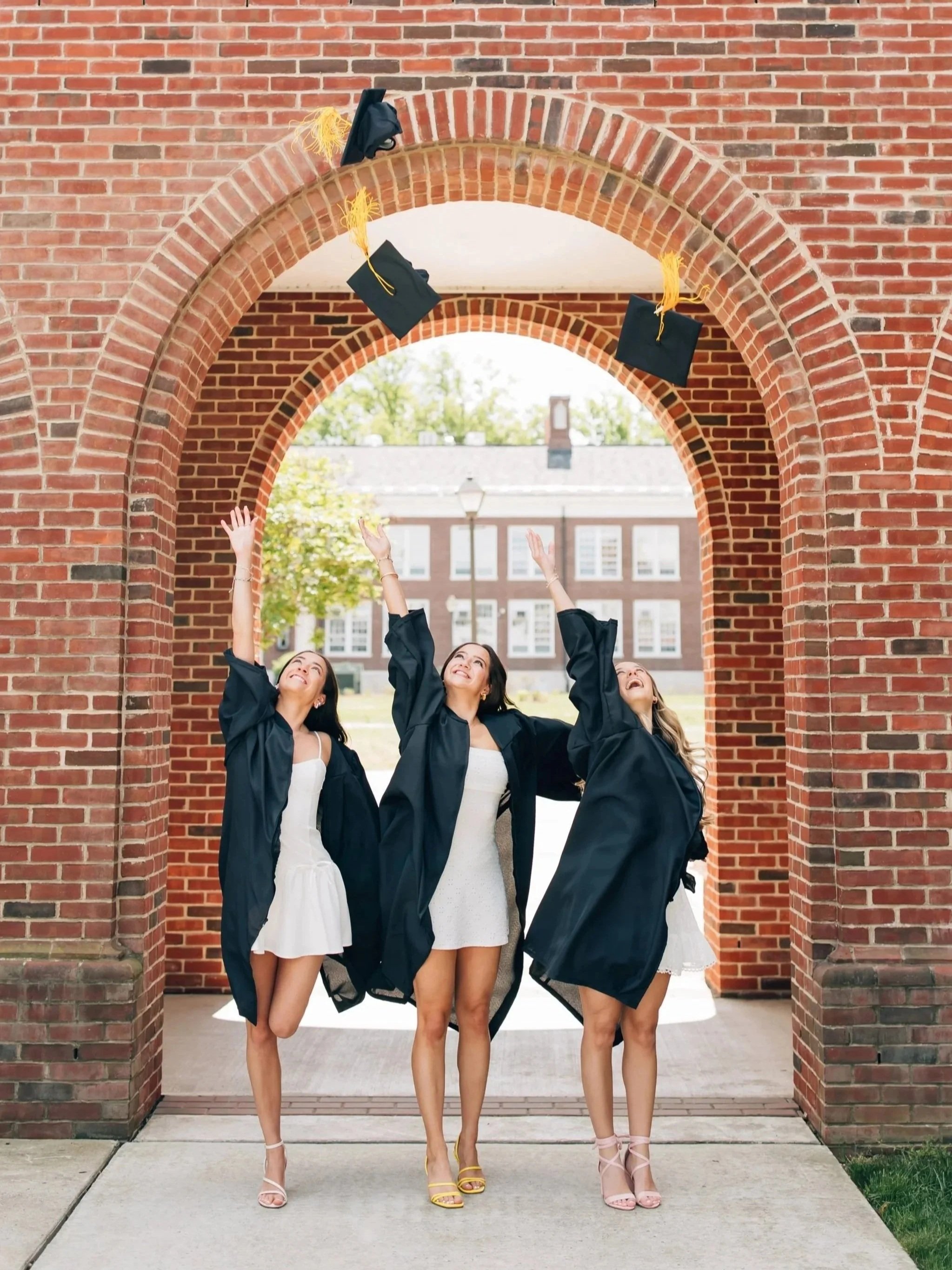 Three women in graduation gowns and dresses celebrating outdoors by throwing their caps in the air under a brick archway.
