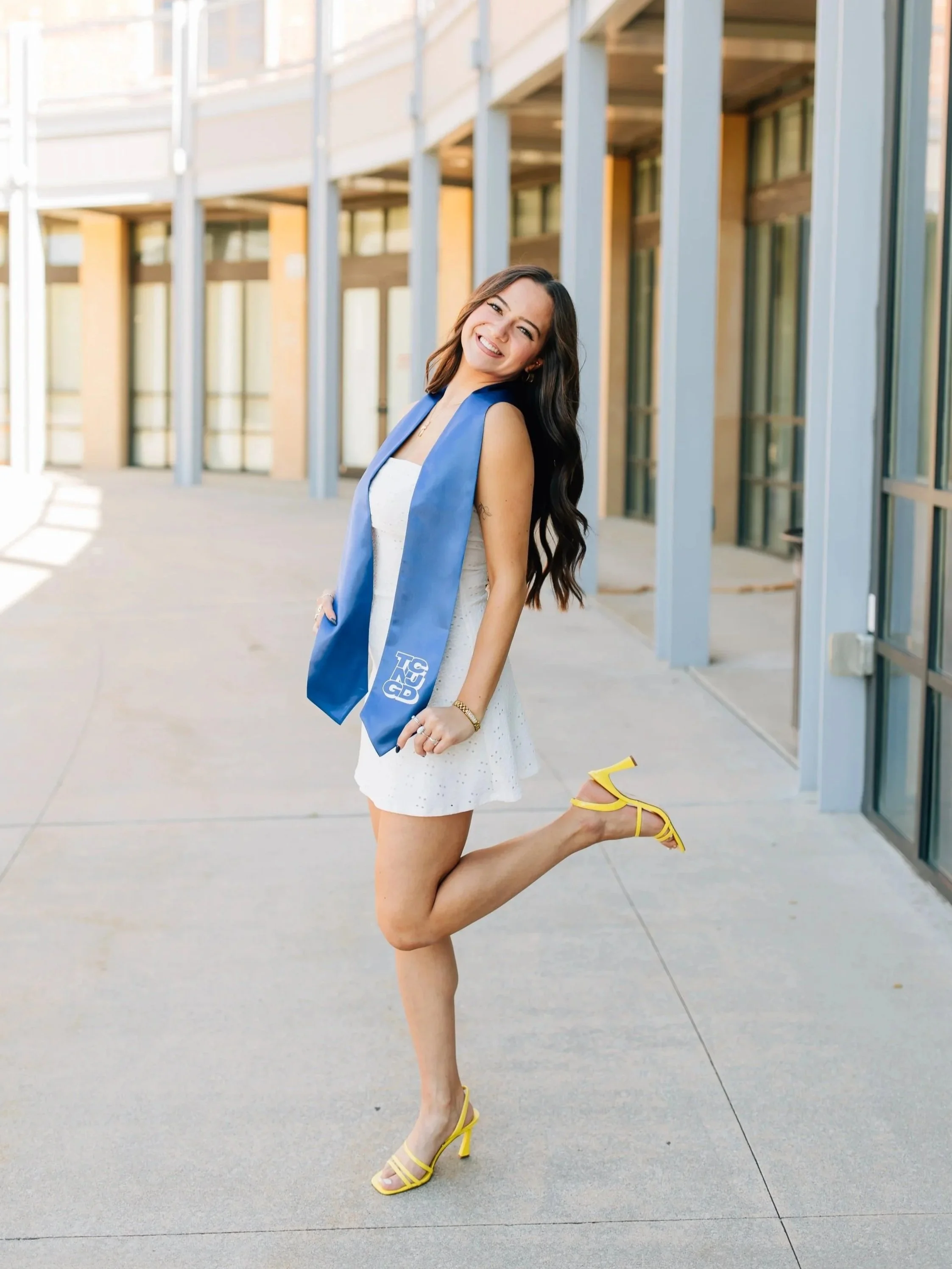 Smiling young woman in white dress and yellow heels posing outdoors in front of modern building.