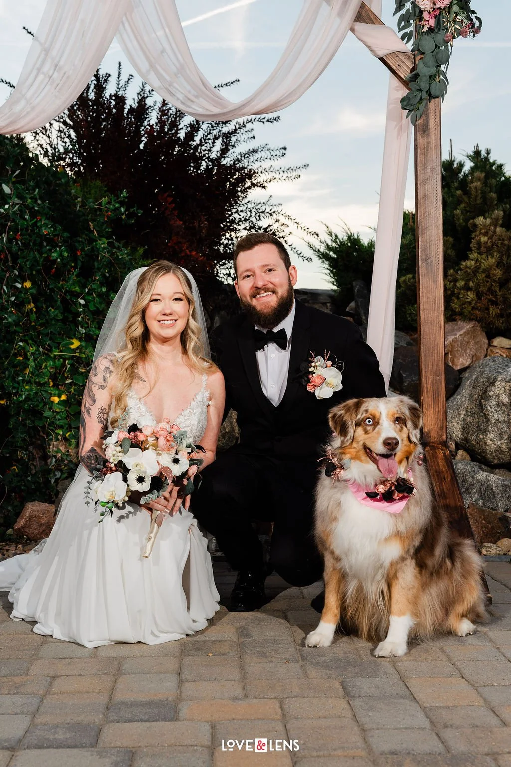 A bride and groom sitting outdoors under a wooden wedding arch with white drapes and floral decorations, smiling with their Australian shepherd dog wearing a pink bandana, on a stone-paved area surrounded by bushes and rocks.