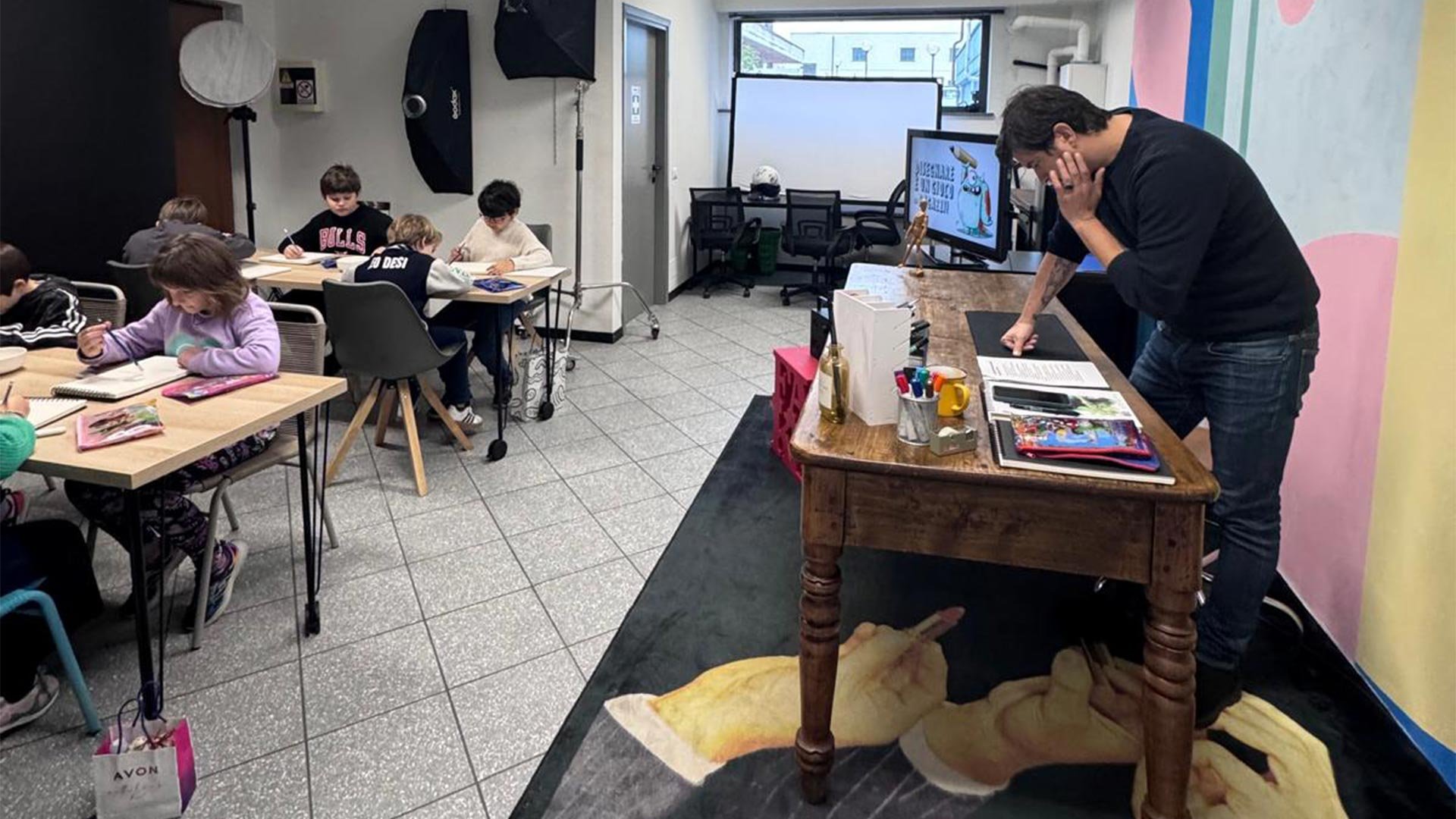 A classroom with children sitting at desks, a teacher at a wooden desk with a digital screen behind him, and a colorful painted wall on the right.