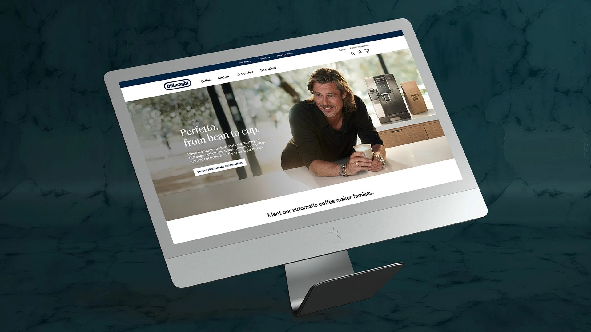 An iMac computer on a dark textured surface displaying the Delonghi website, featuring a man with long hair and a beard holding a coffee mug in a modern kitchen.