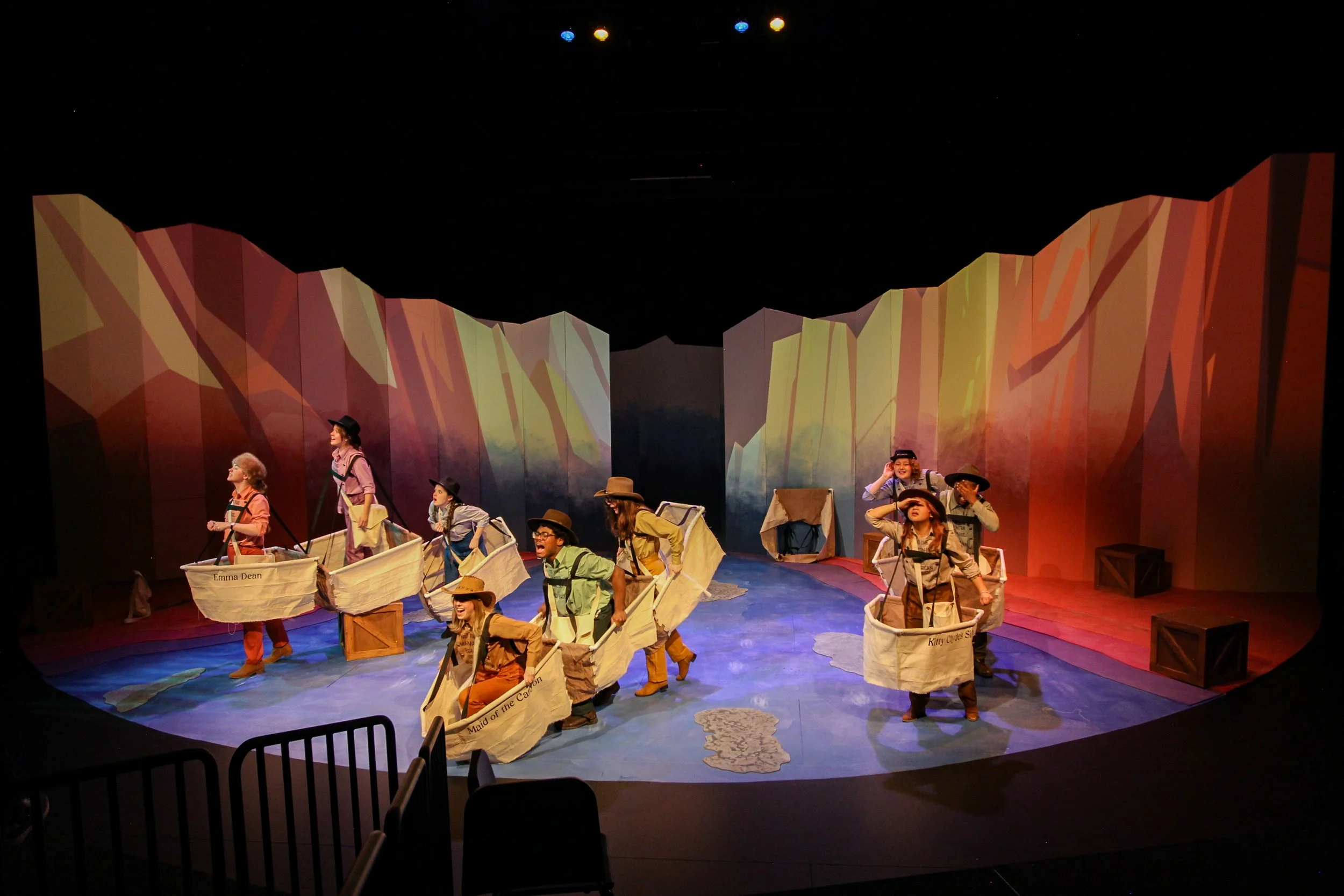 Children performing a play on stage, dressed as explorers with paper boats, in front of colorful mountain-like backdrop.