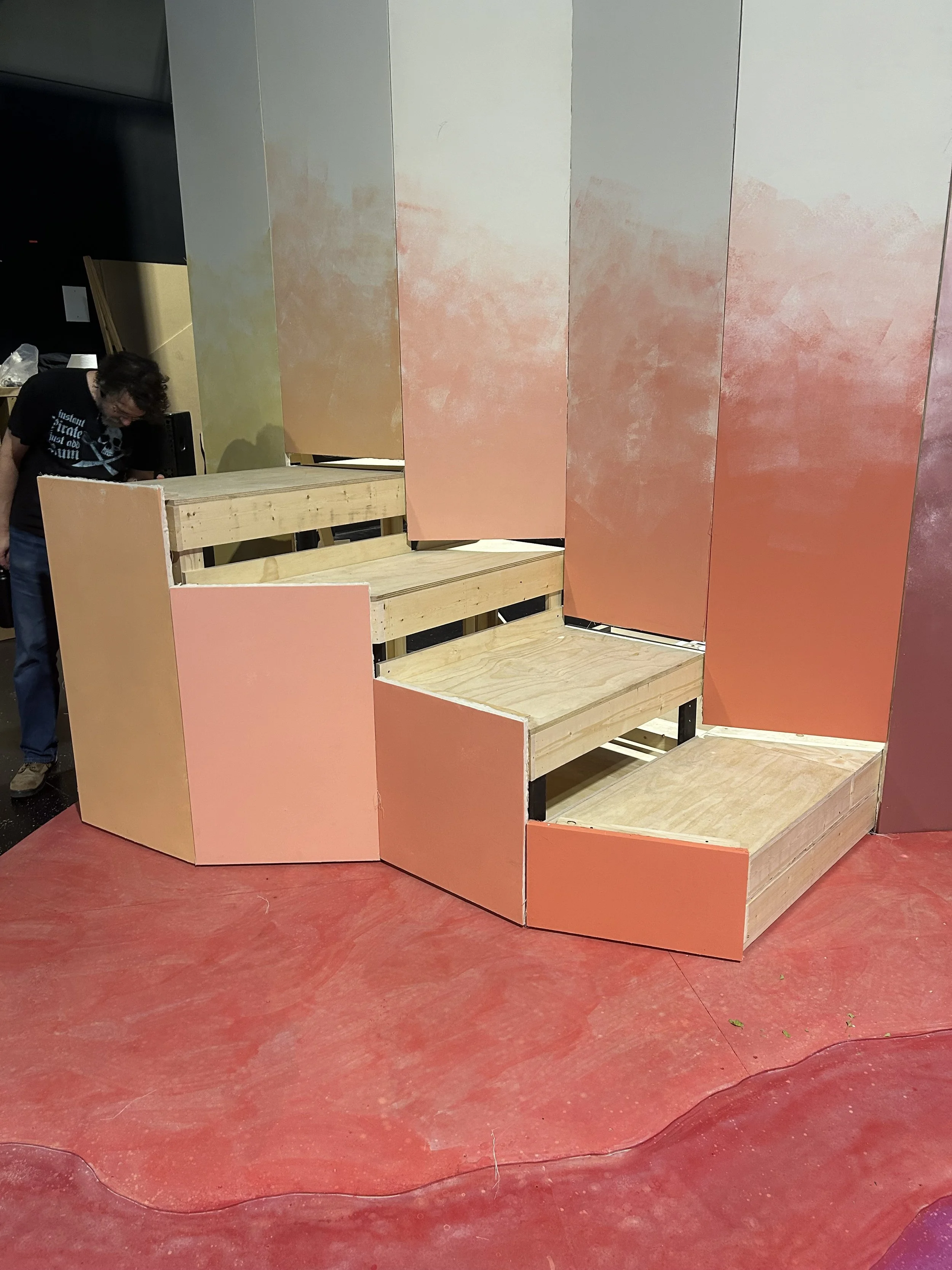 A partially constructed stage with pink panels and wooden steps, with a person working on the left side.
