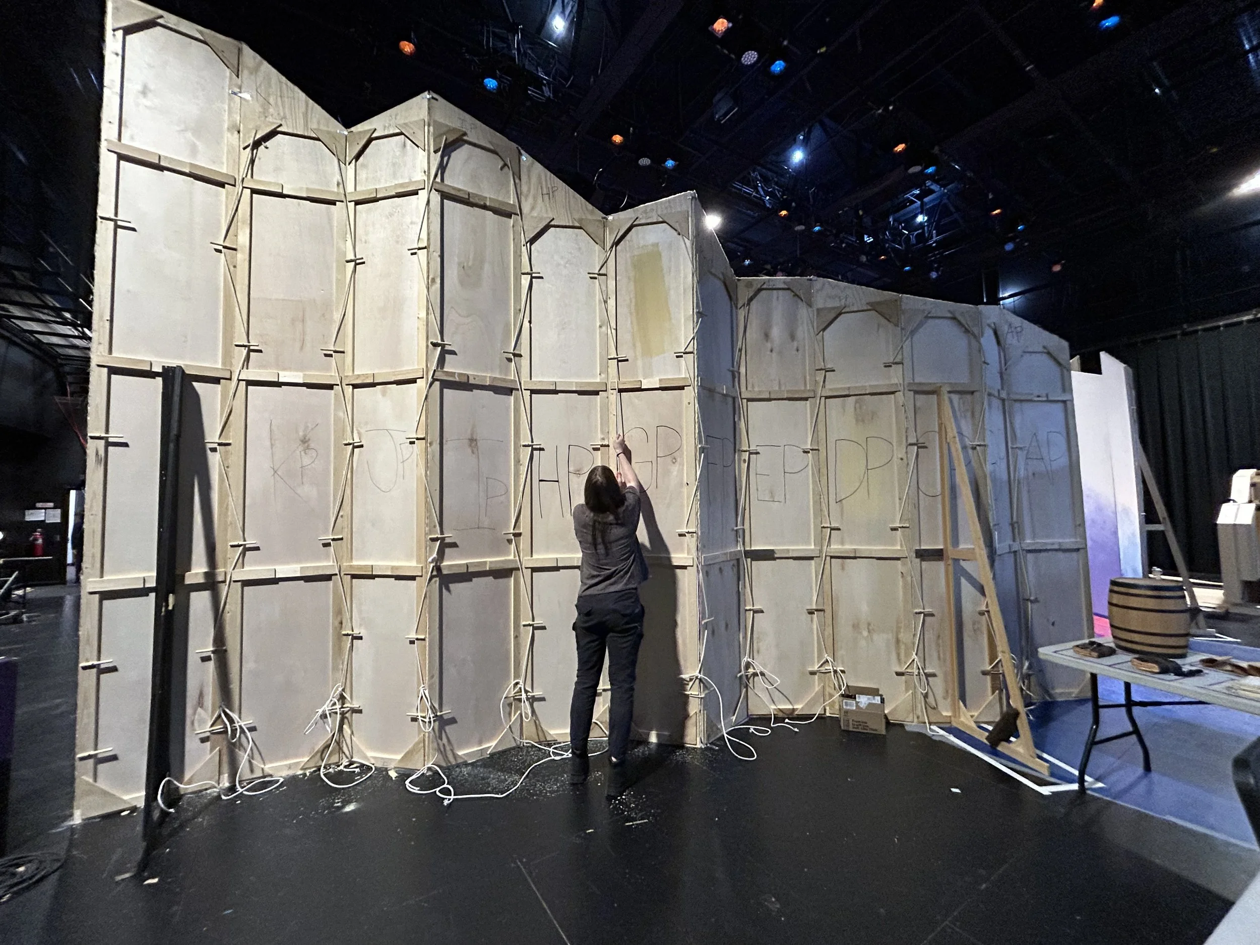A woman working on a large wooden structure with vertical and horizontal supports, possibly creating a set or backdrop, in a dimly lit room with stage equipment and tables nearby.