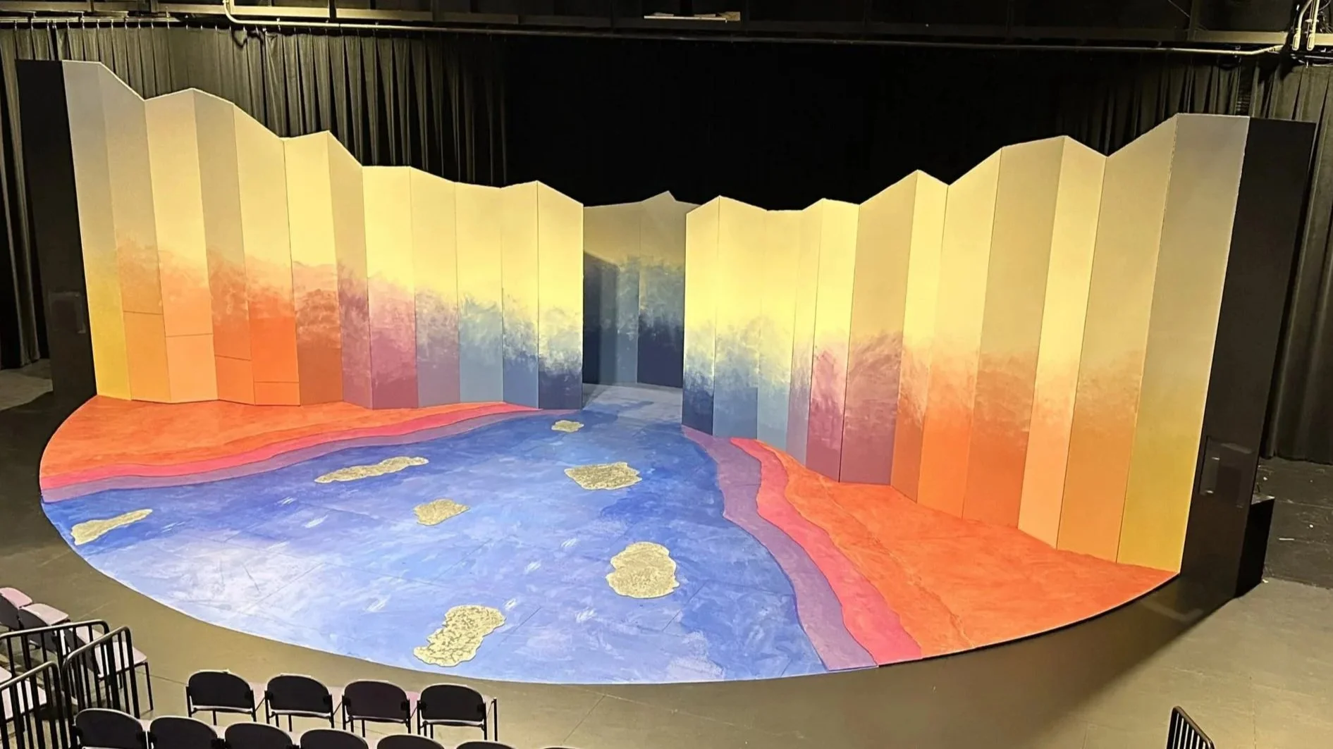 Colorful stage set with a large, curved backdrop featuring a landscape of trees and sky, with a painted floor resembling a river and land, in a theater with empty black chairs.