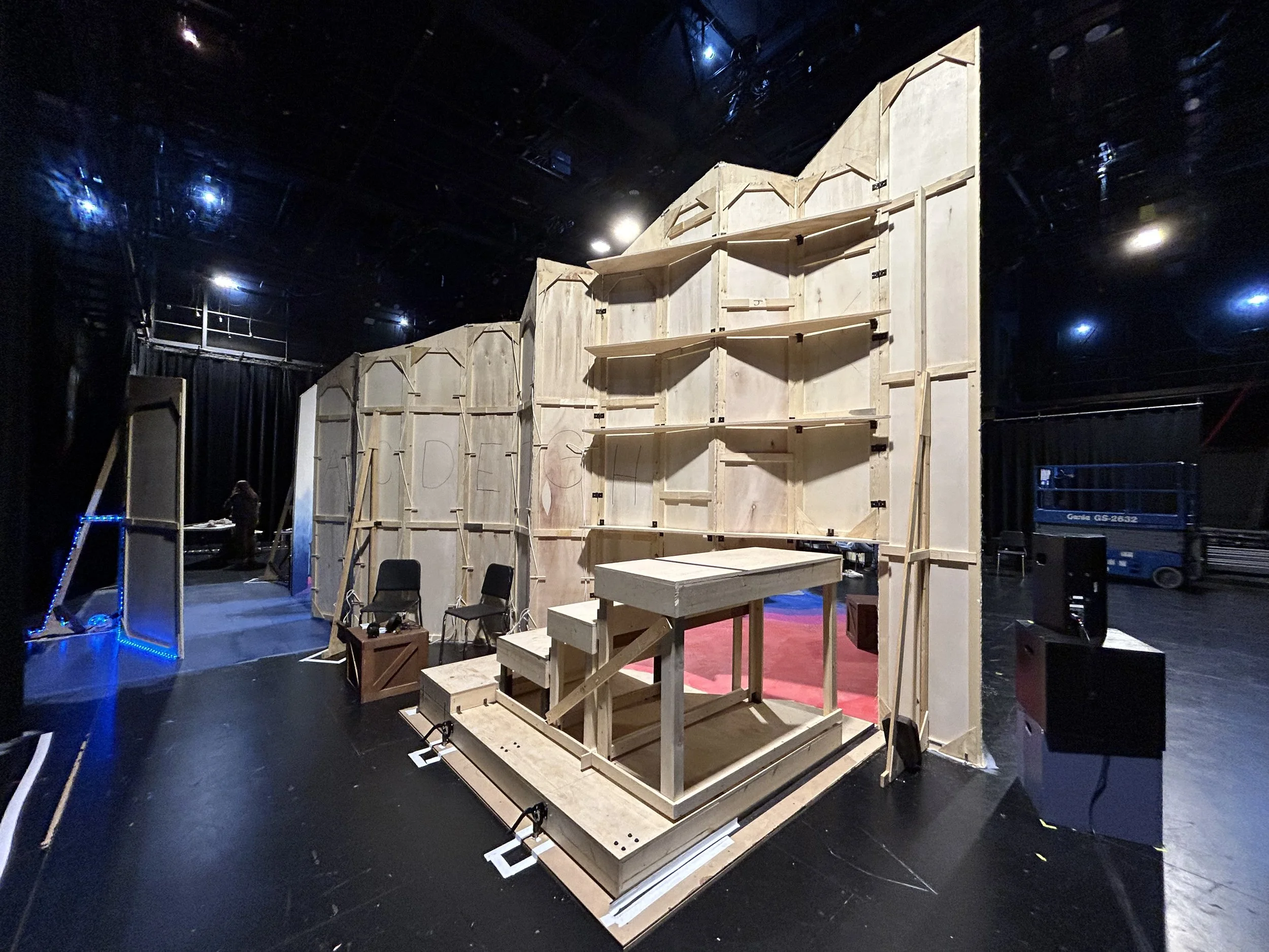 The image shows a theatrical stage set under construction with wooden panels and scaffolding, and black curtains in the background.