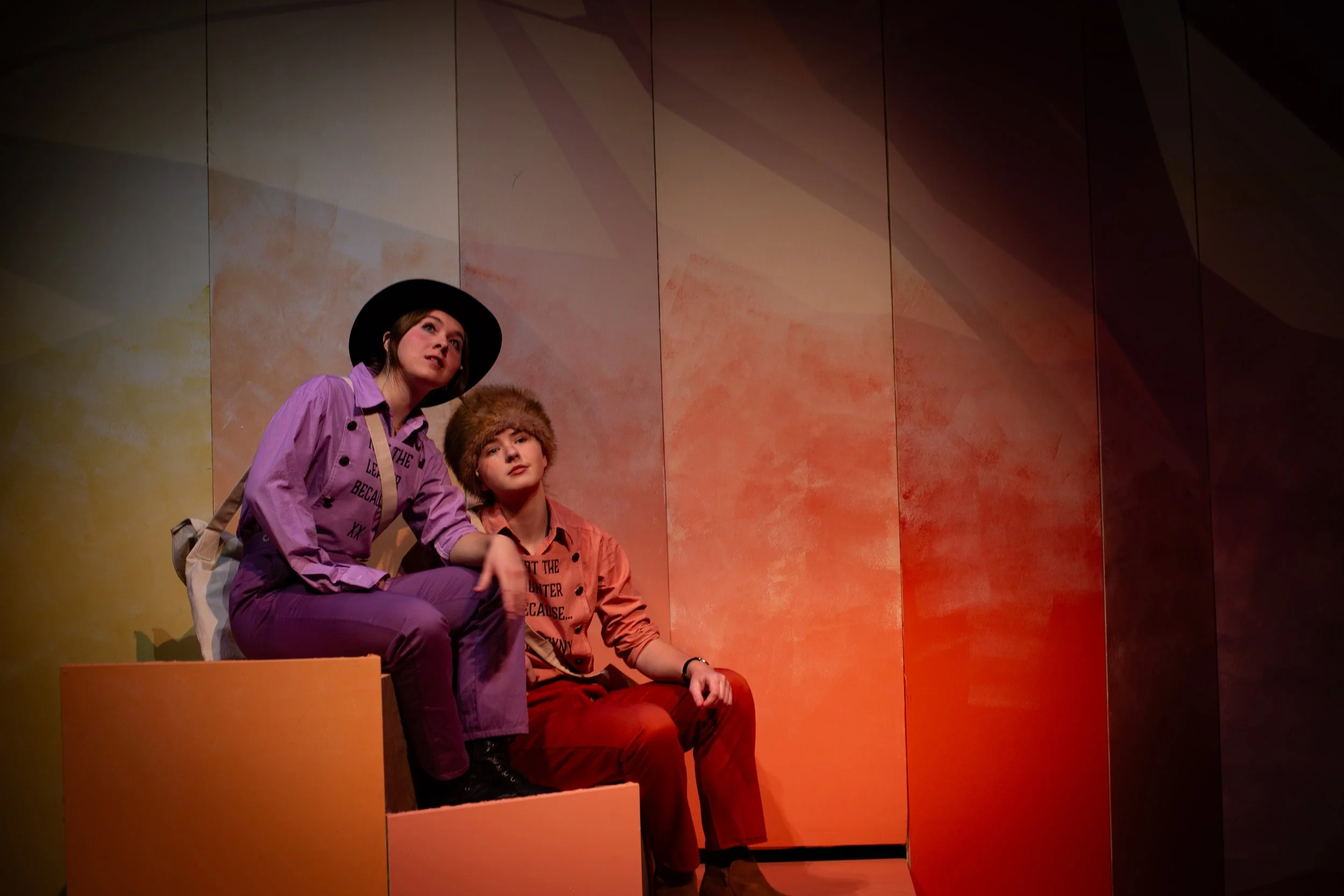 Two young people sitting on a pink platform, with a colorful gallery wall behind them, one wearing a purple jacket and black hat, the other wearing a tan hat and red pants, in a museum or art gallery.