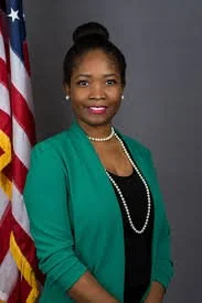 A Junie Joseph standing in front of an American flag, wearing a green blazer and pearl necklace, smiling at the camera.