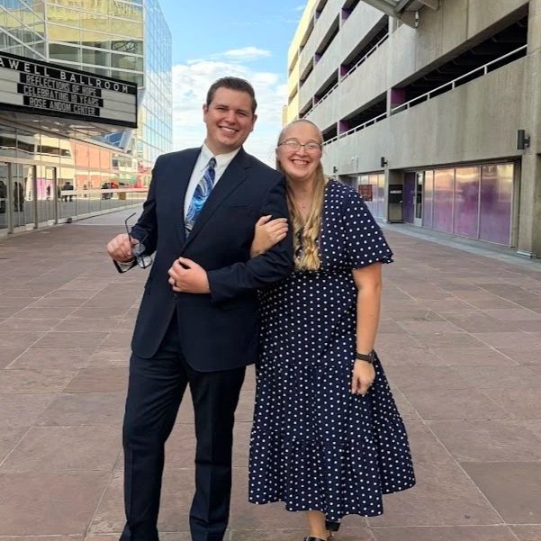 A man in a suit and a woman in a polka dot dress pose together outside a modern building, smiling and holding hands.