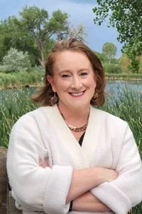 A woman with shoulder-length red hair smiling outdoors by a pond, wearing a white sweater and a necklace, with greenery and trees in the background.