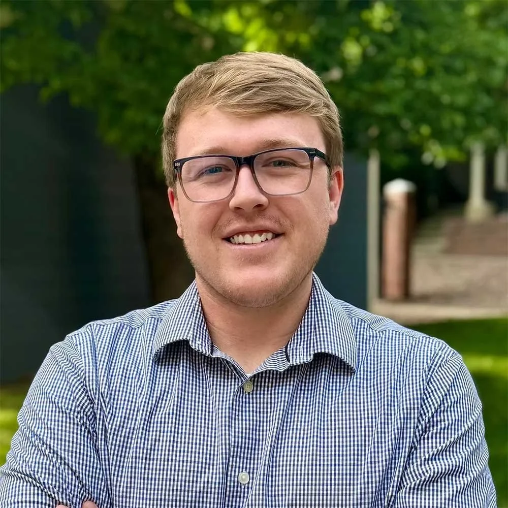 Young man Jack Johnson with glasses smiling outdoors with trees and stairs in the background.